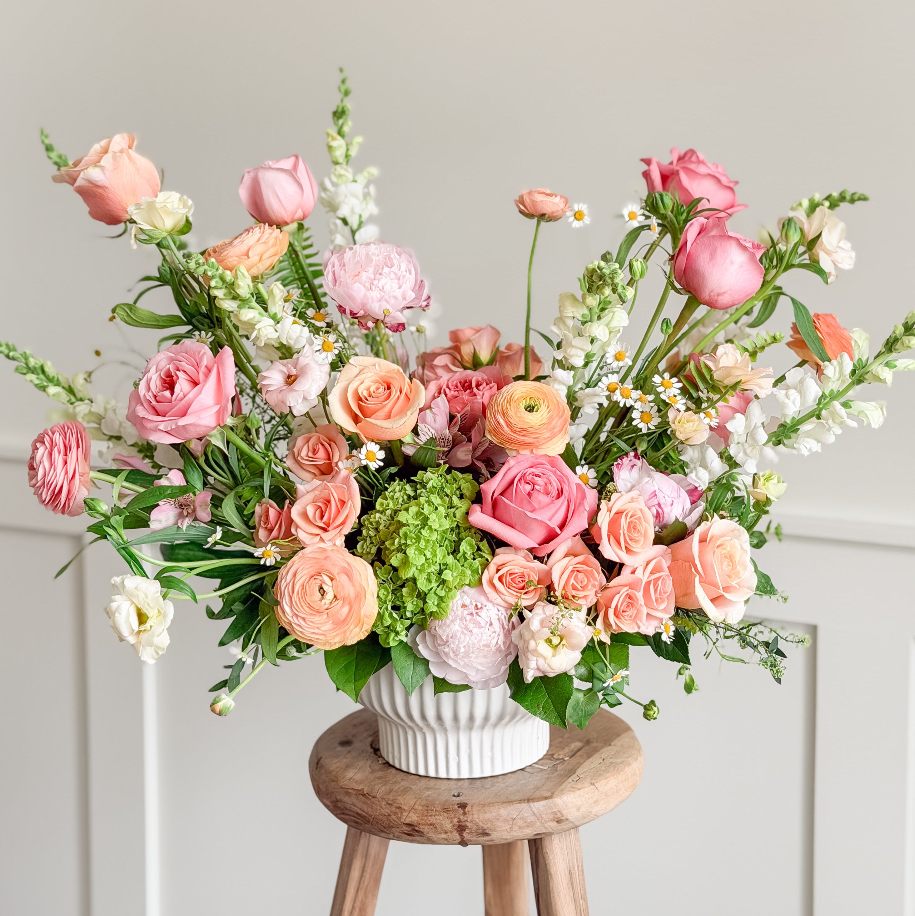 Bouquet of pink and peach flowers in a white vase on a wooden stool against a light gray wall from Lovebird Flowers, London Ontario's best florist.