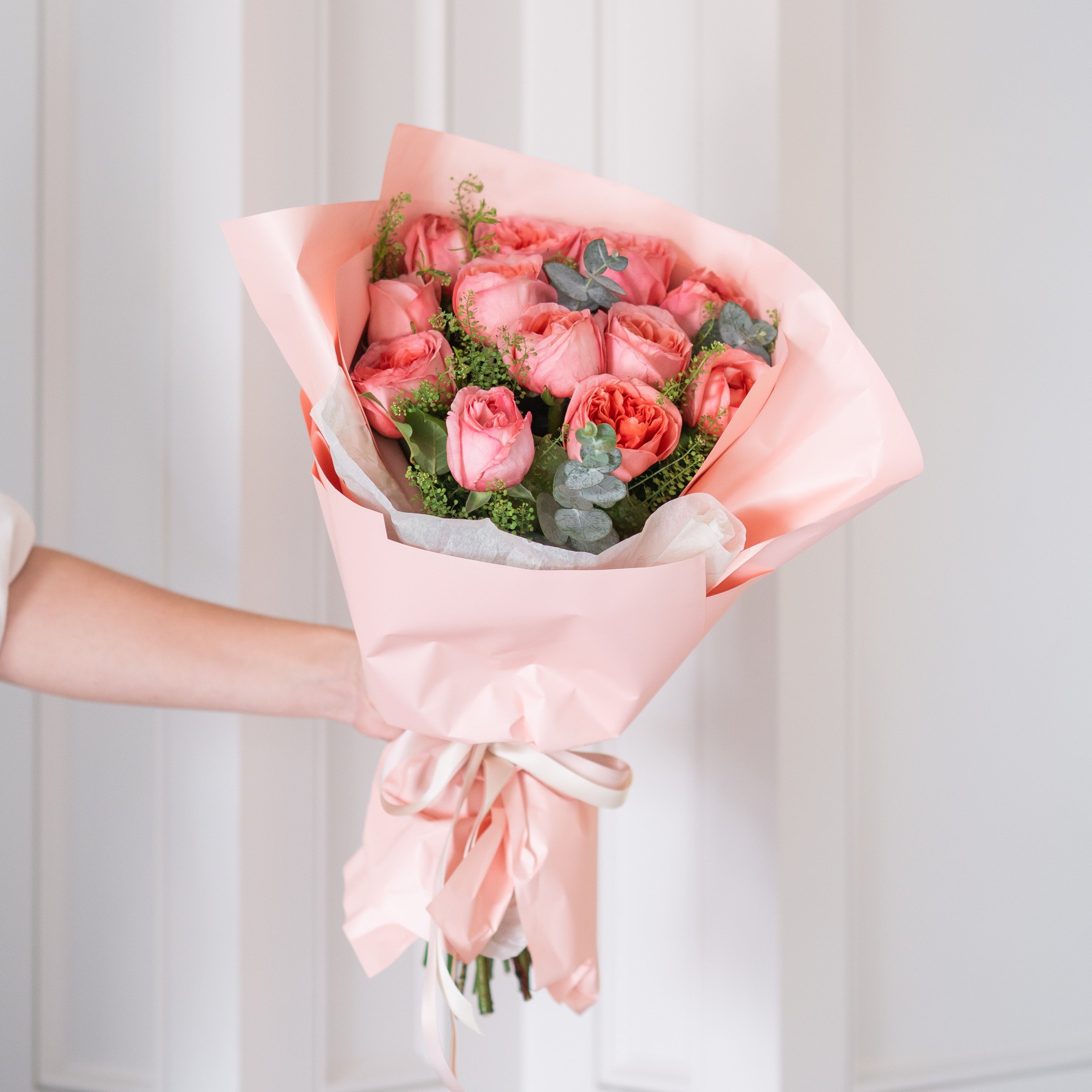 Bouquet of pink flowers with greenery in a white container, wrapped in pink paper and ribbon, held by a person against a light gray background.