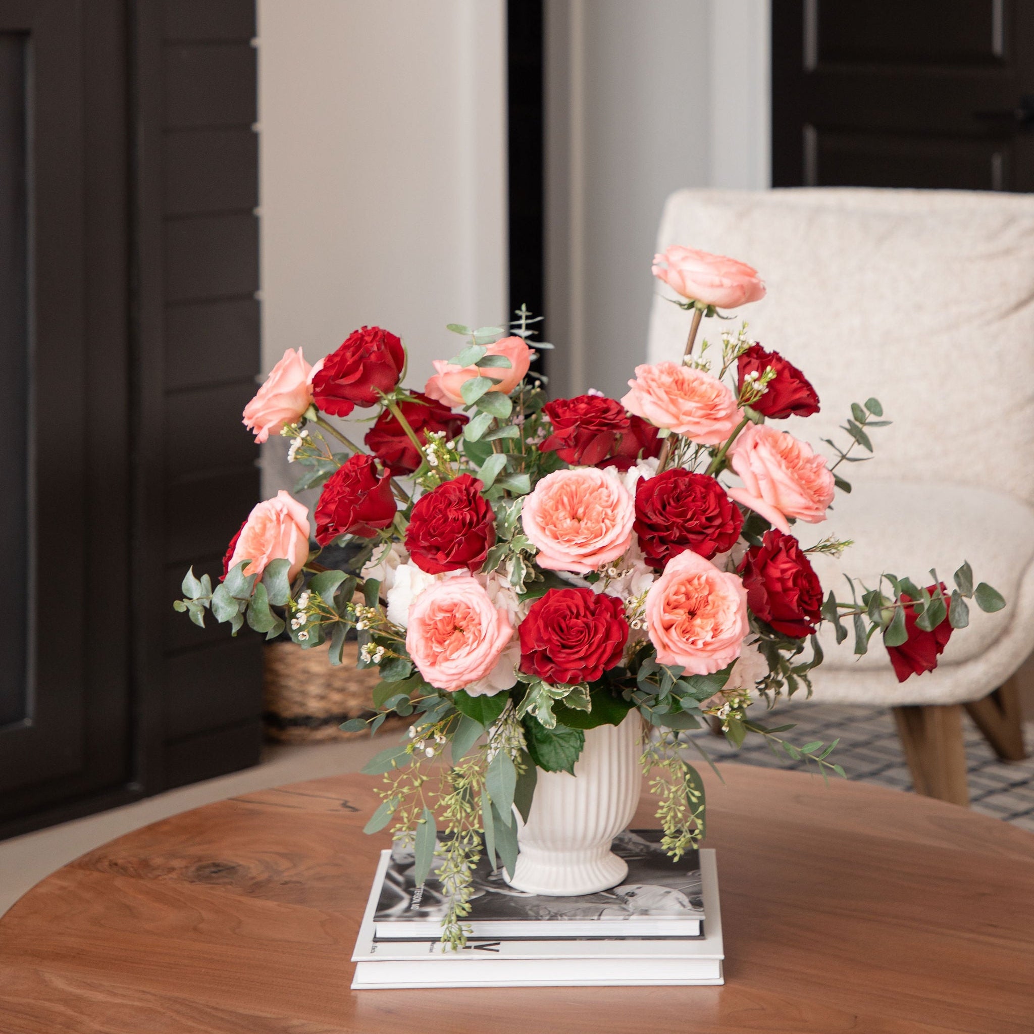 Bouquet of red and pink flowers in a vase on a wooden table with a white chair and fireplace in the background.