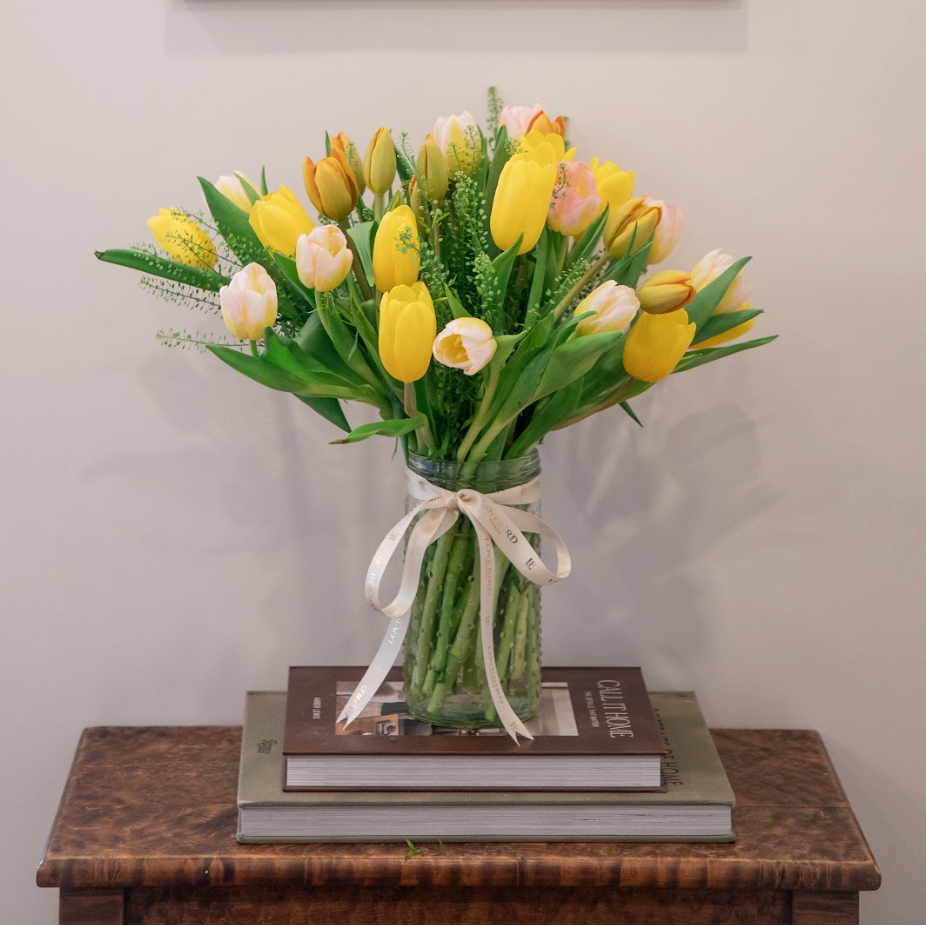 Bouquet of yellow and white tulips in a glass vase on a wooden table with a framed artwork in the background.