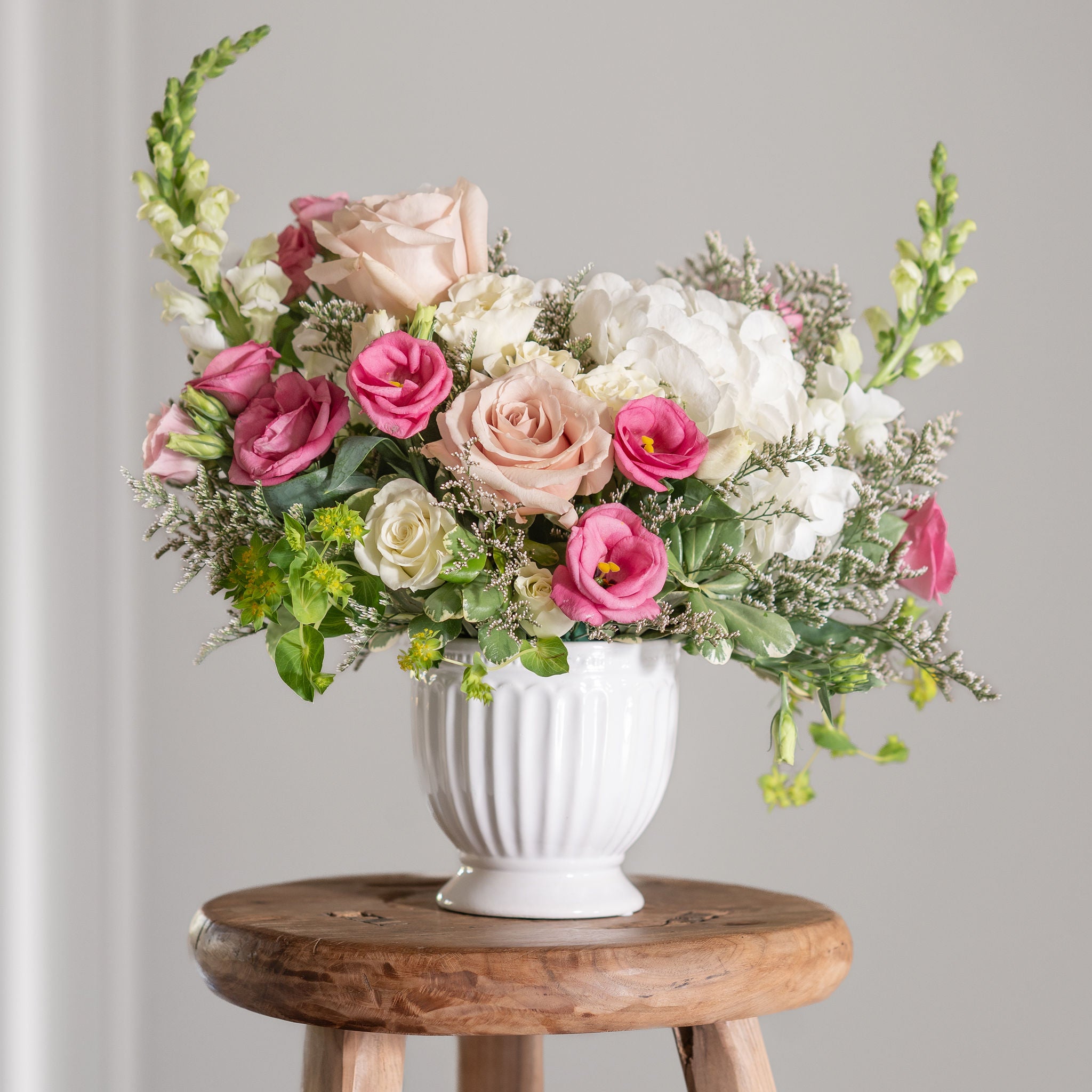 Flower arrangement with pink lisianthus, quicksand roses, hydrangea, limonium, snap dragons in white footed compote