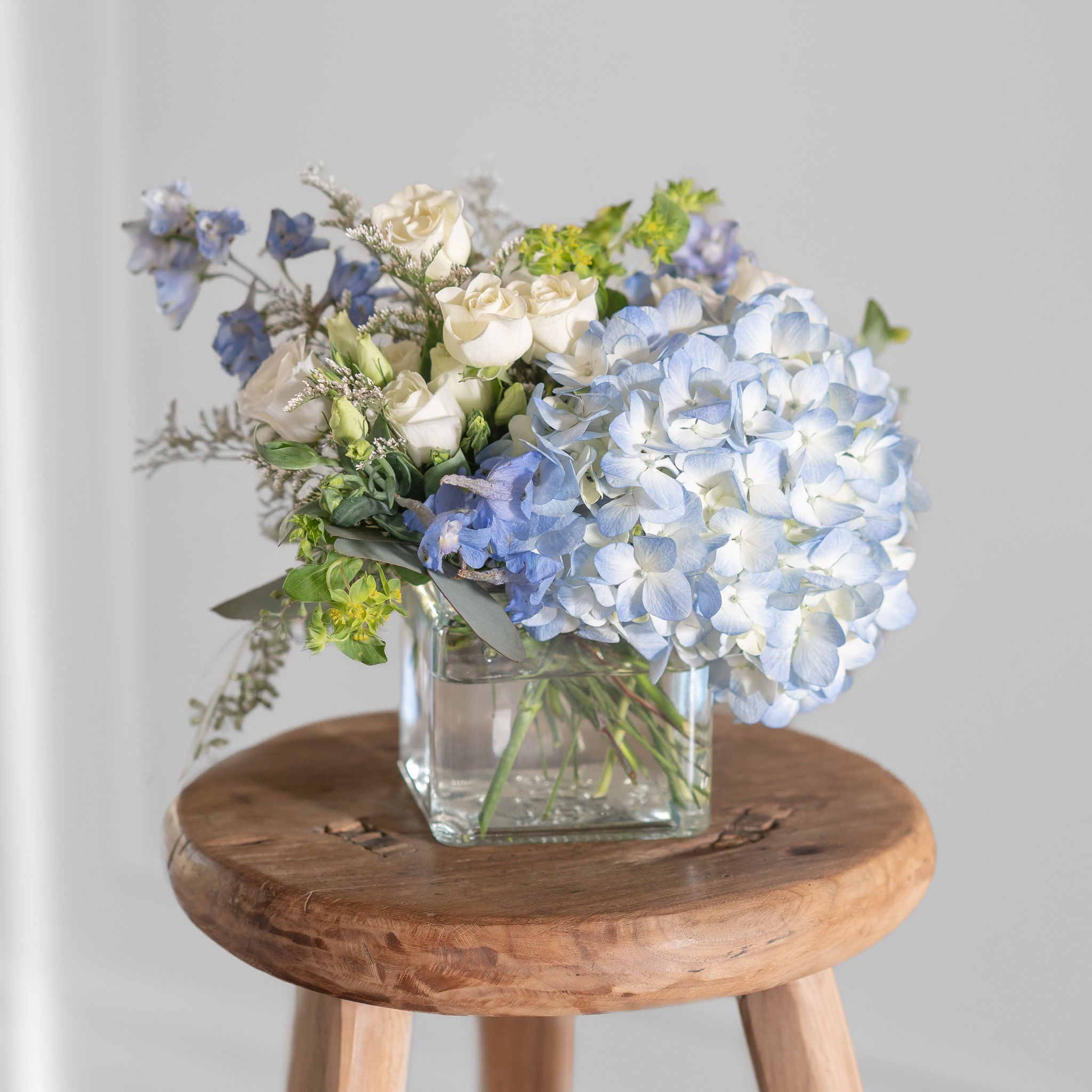 blue hydrangea, blue delphinium, spray roses, liisianthus arrangement in a clear cube vase on a wood stool