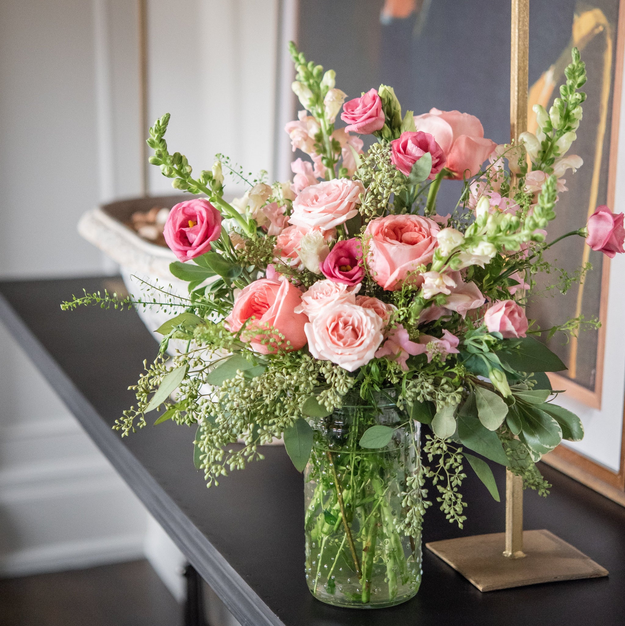 Bouquet of pink roses, pink lisianthus, pink snapdragons and seeded eucalyptus flowers in a clear vase on a dark surface.