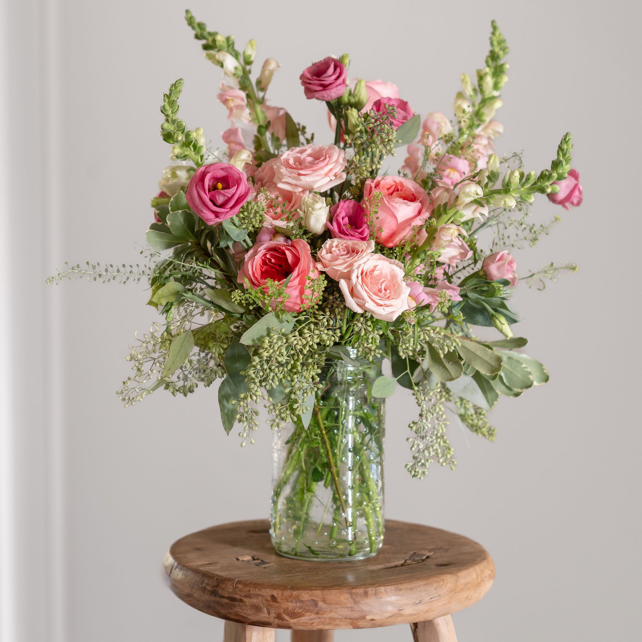Bouquet of pink garden roses, pink spray roses, pink lisianthus and snapdragons in a clear vase on a wooden stool with a white background