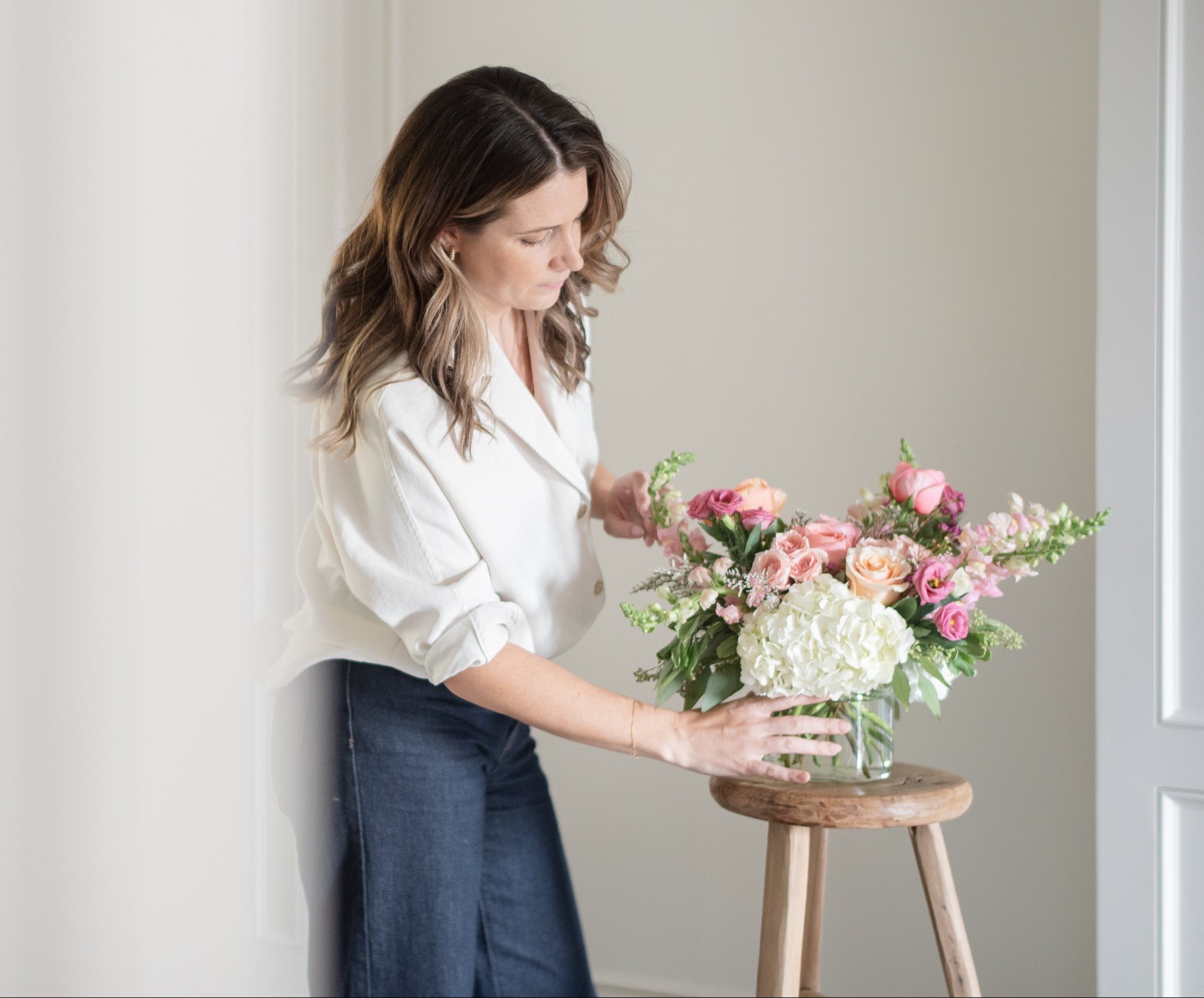 woman arranging white and pink flower arrangement on a stool with pink and peach spray roses, pink snap dragons and white hydrangea