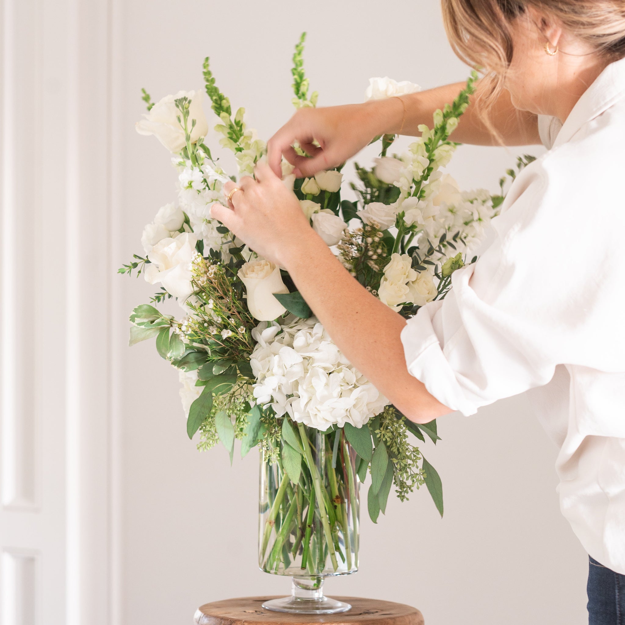 woman arranging large white flower arrangement wiht snap dragons, white roses, hydrangeas in a pedestal vase for sympathy