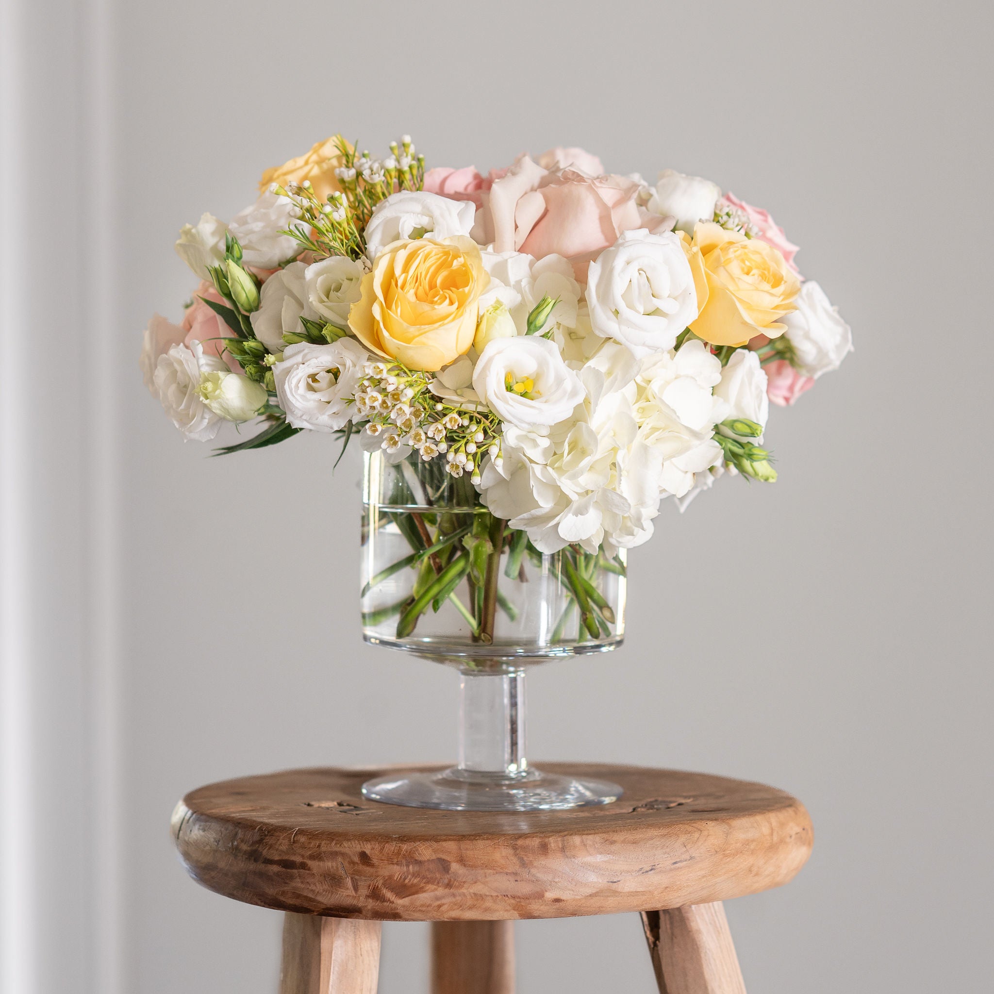 Soft yellow buttercup roses, quicksand roses, white hydrangea, white lisianthus and wax flower in a premium pedestal vase.