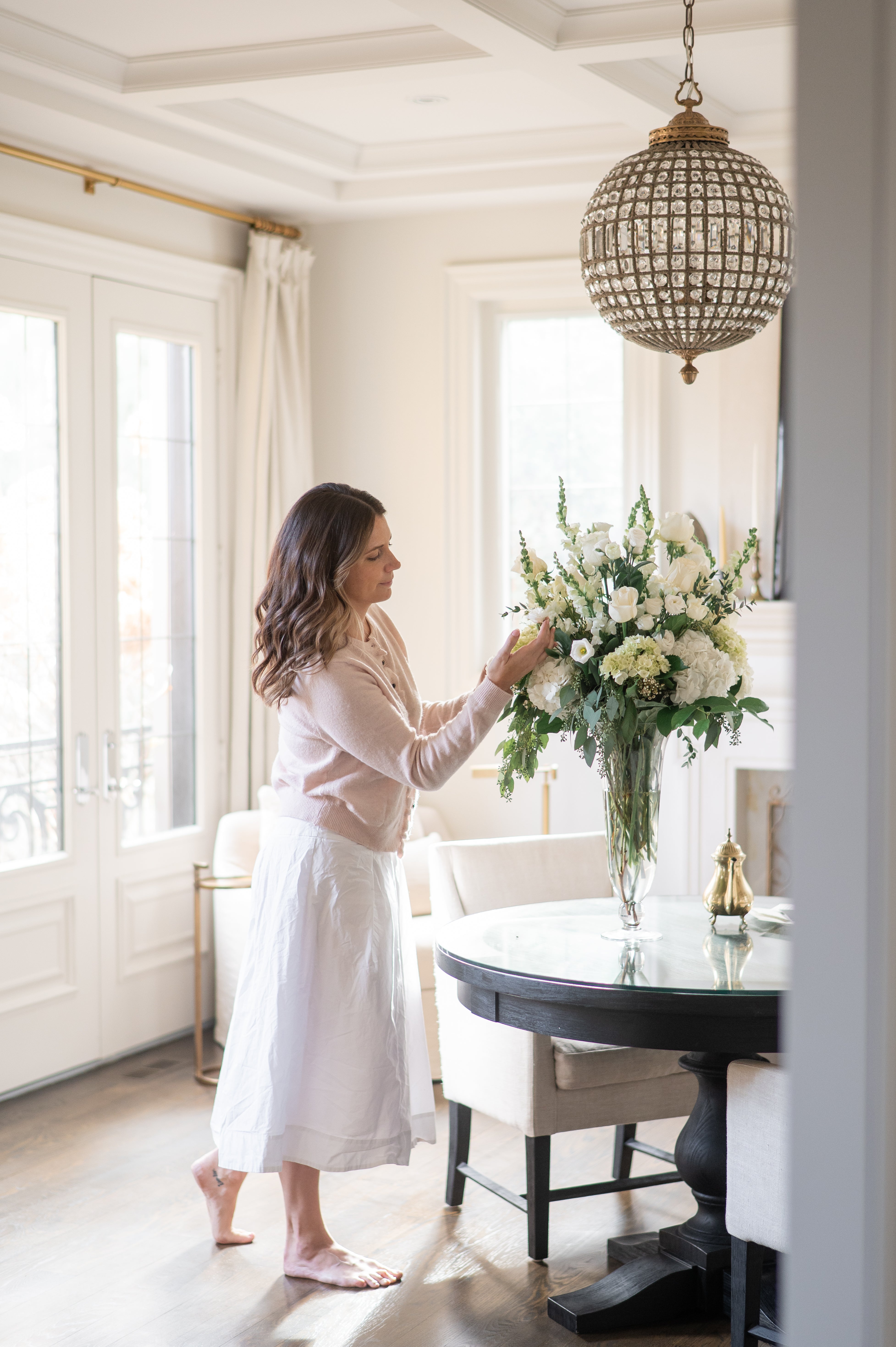 Woman arranging a grand pedestal vase of white sympathy flowers featuring roses, snap dragons, hydrangeas in a bright, elegant room with large windows and a chandelier.