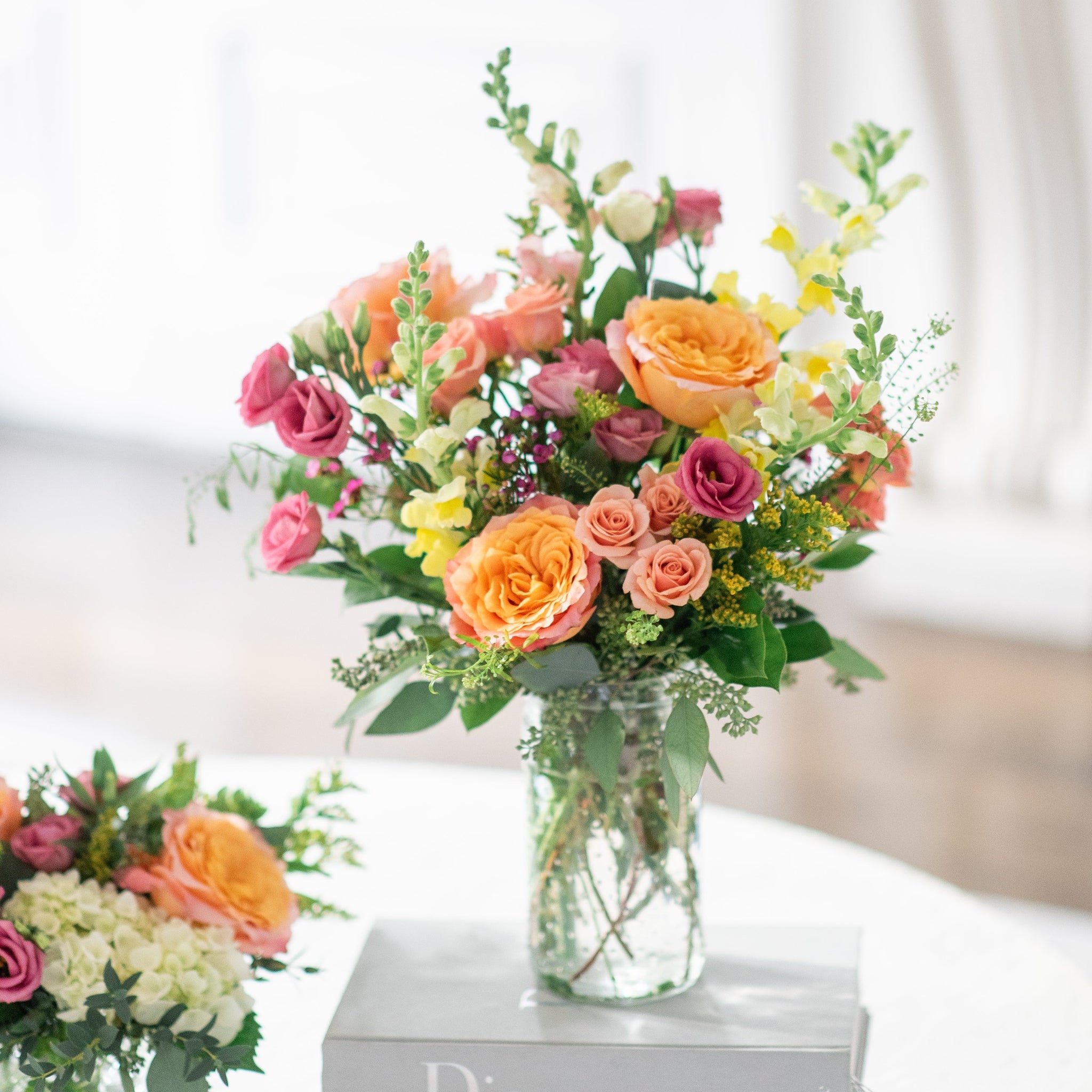 Colorful flower arrangement with free spirit roses, yellow snap dragons, golden rod, pink lisianthus in a vase on top of a Chanel book with a blurred background