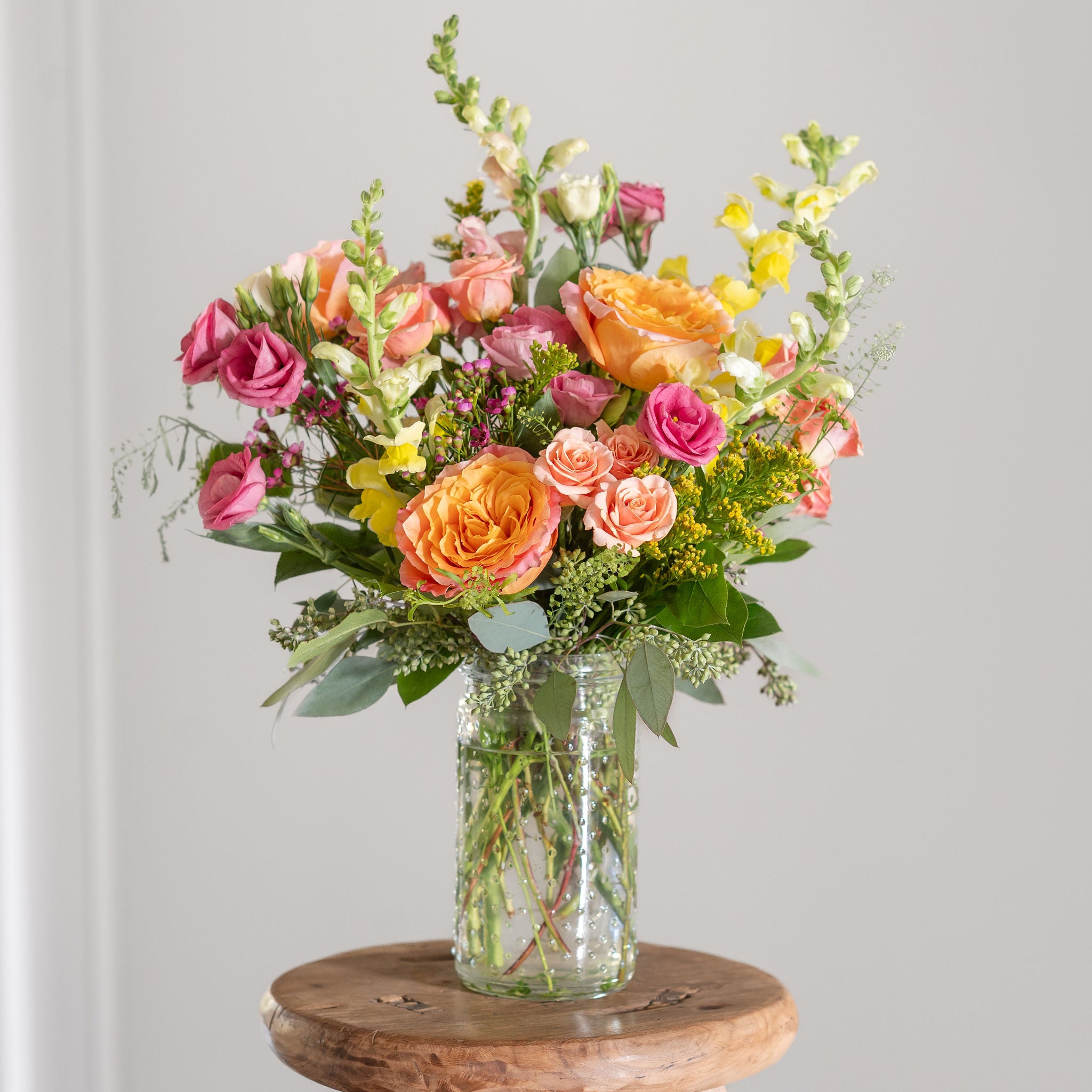 Bright orange, peach, pink and yellow rsoes, snap dragons, lisianthus, golden rod, and seeded eucalyptus in a clear vase on a wooden stand against a gray background