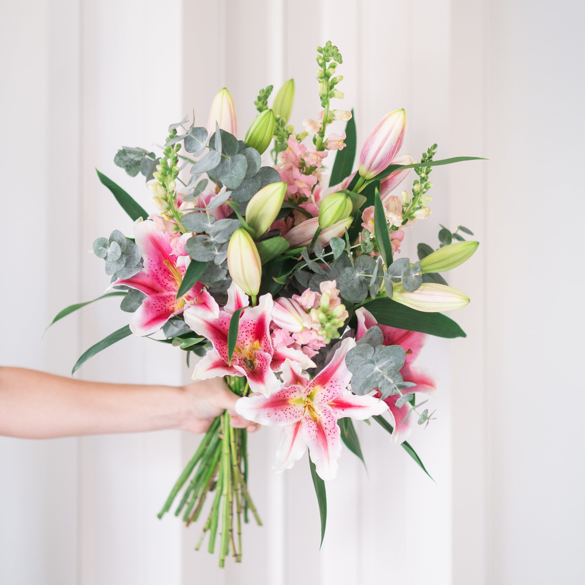 Bouquet of pink lilies and greenery held by a person against a white curtain background