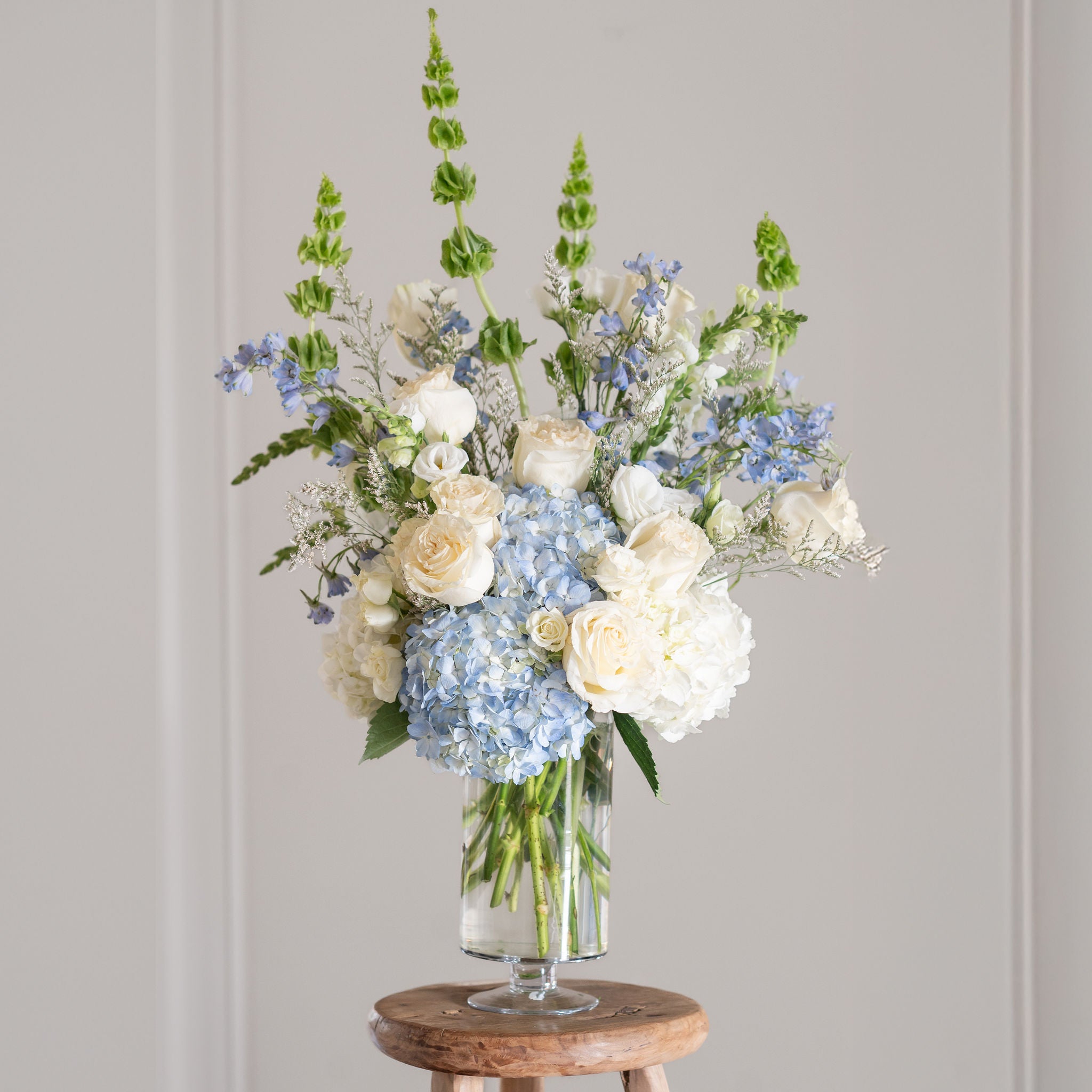 white and blue tall arrangement with bells of ireland, roses and hydrangea in a premium pedestal vase