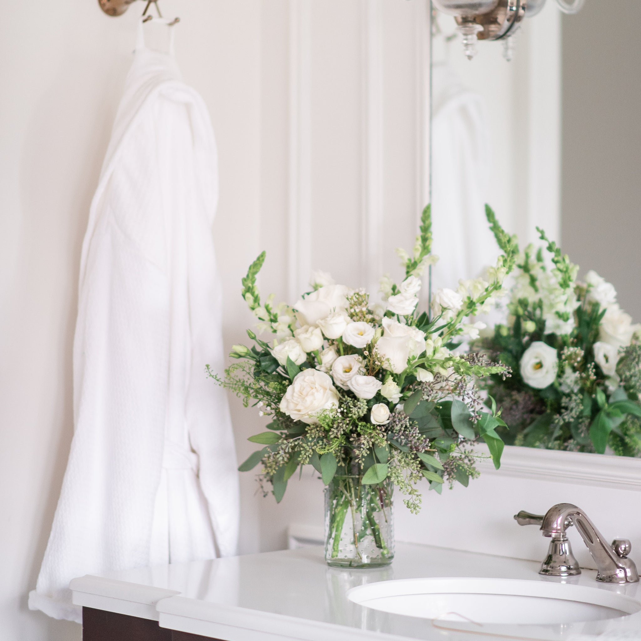 Bathroom with a sink, mirror, and white snapdragon, white rose and seeded eucalyptus floral arrangement on a white countertop.