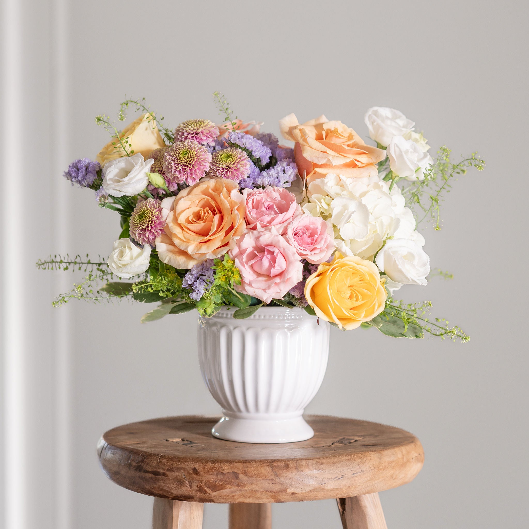 Pastel floral arrangement with hydrangea, pink and peach roses, purple statice, in a white vase on a wooden stool with a gray background