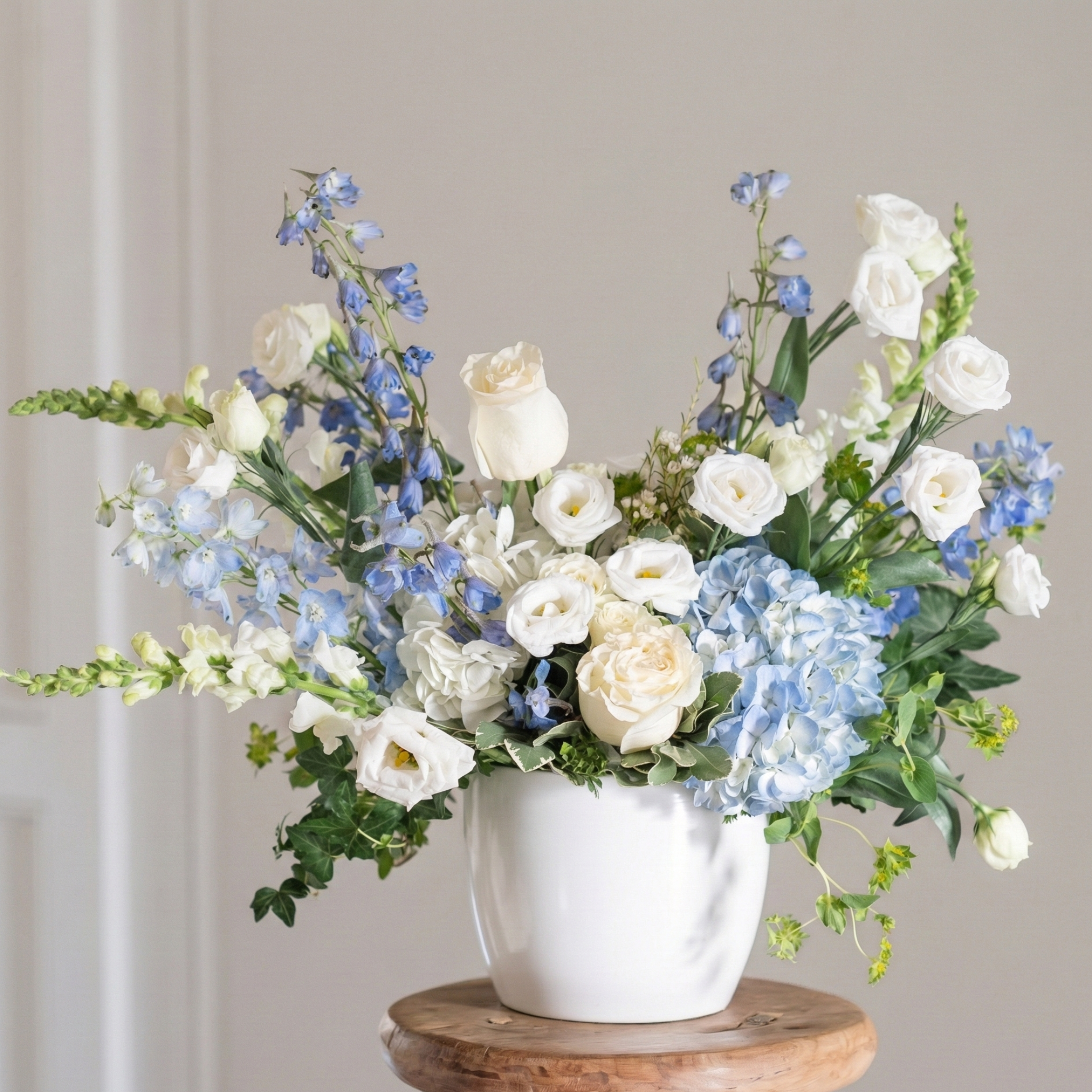 Floral arrangement with white and blue flowers in a white vase on a wooden stool against a neutral background
