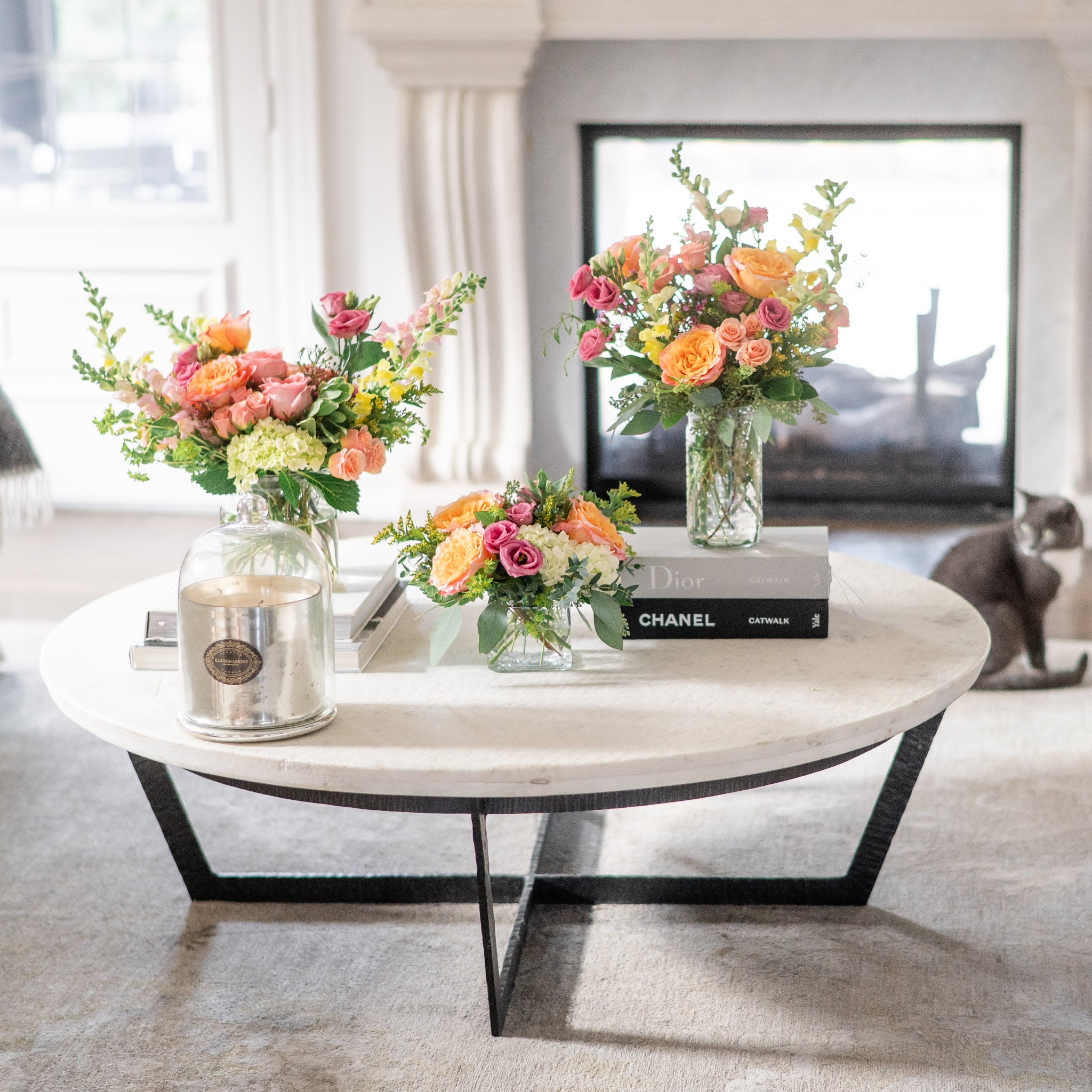 Living room with a coffee table featuring thee bright floral arrangements and books, with a cat sitting on the floor.