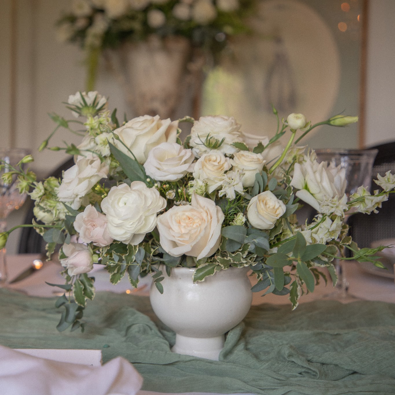 White compote wedding centrpiece with white roses, ranunculus, delphinium, and greenery on a green tablecloth