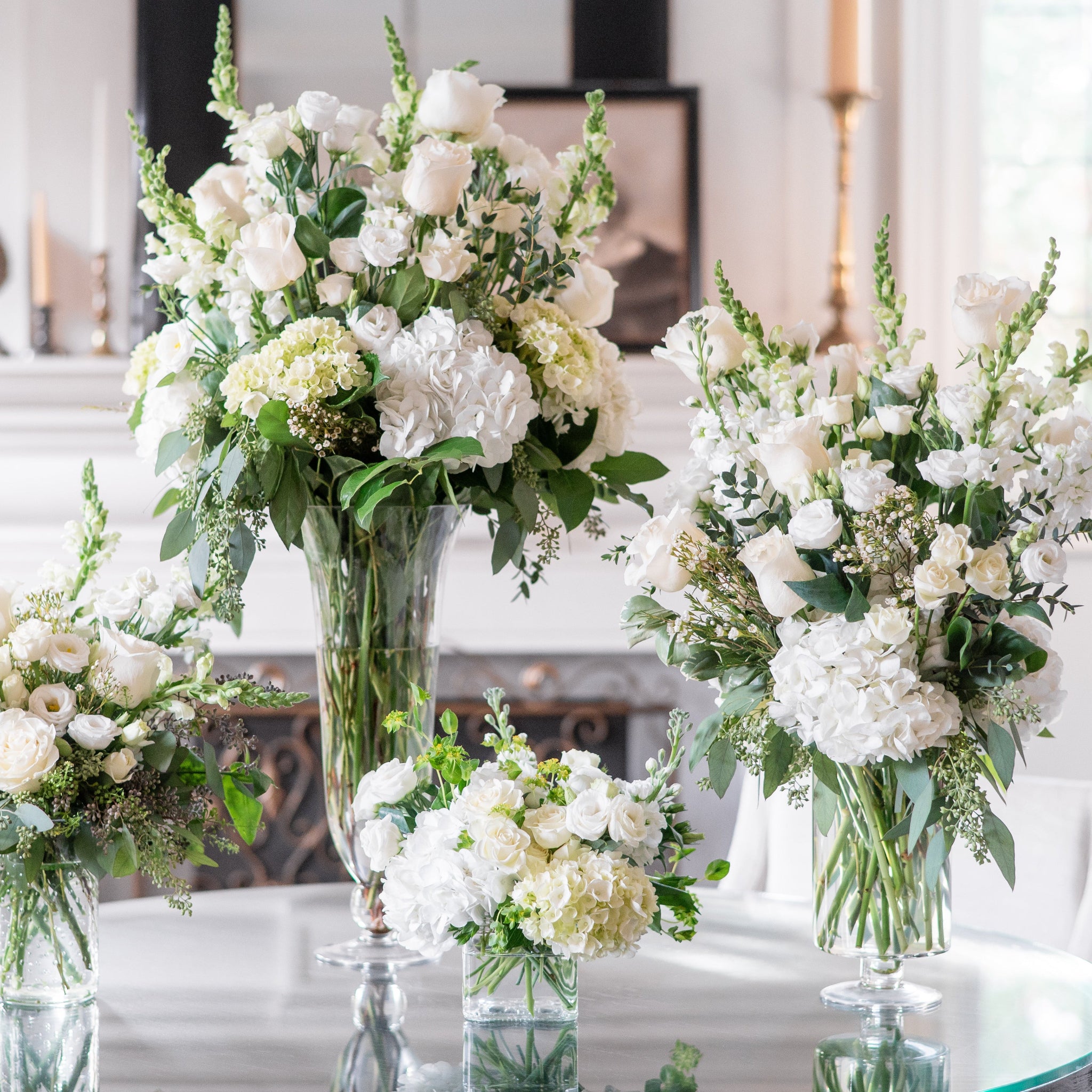 Four white floral arrangements of various sizes on a round table in a bright room.
