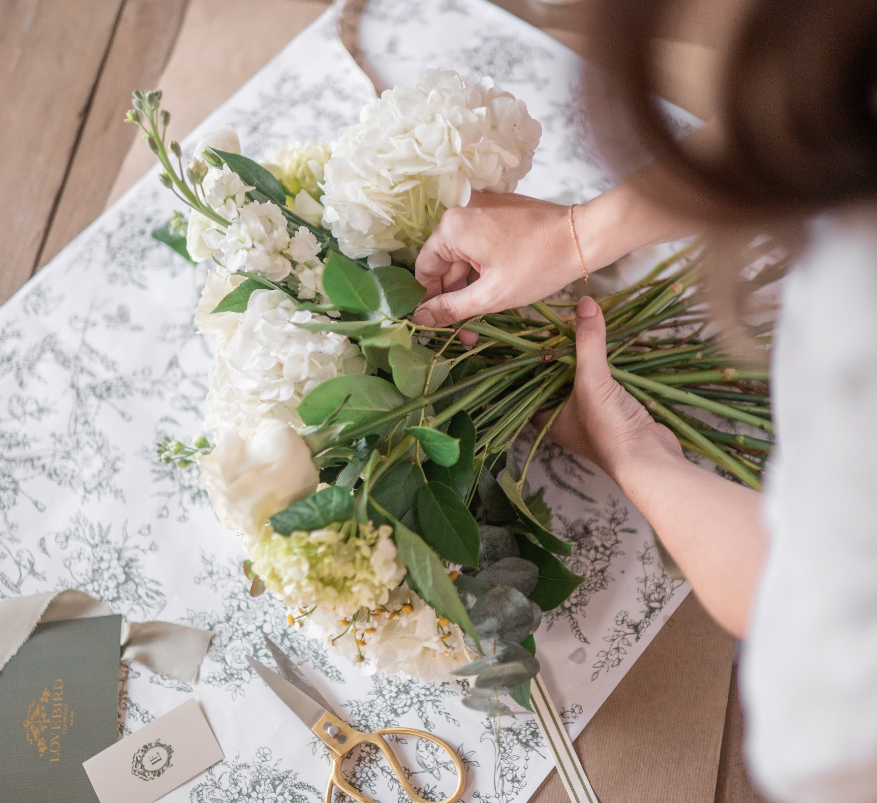 woman laying hydrangea, white roses and snap dragon flowers into a bouquet on a peice of toile wrapping paper with scissors and Lovebird Flowers cards