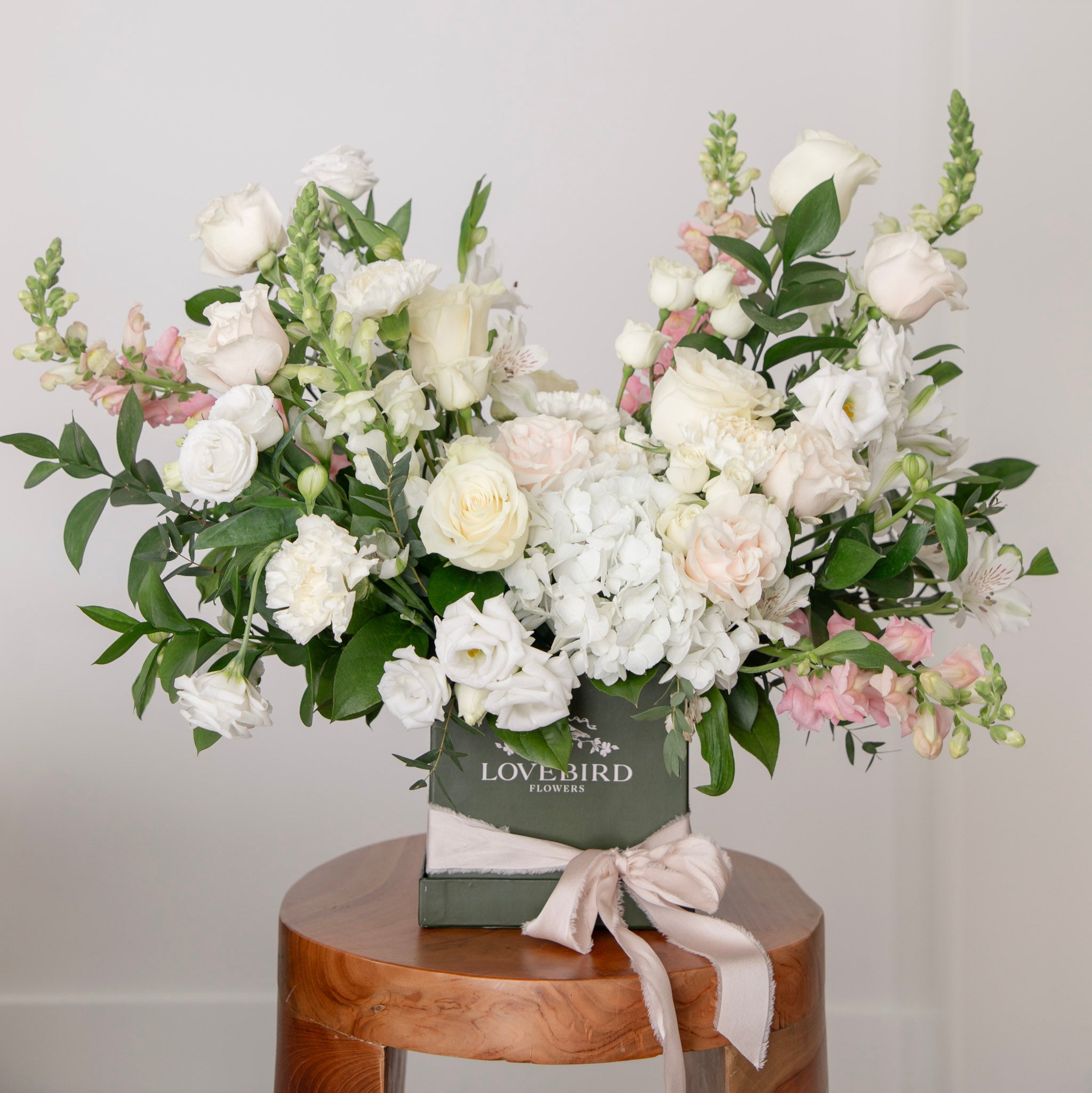 Always & Forever luxury white and blush floral arrangement with hydrangeas and roses in a signature green Lovebird Flowers hat box, displayed on a wooden stool