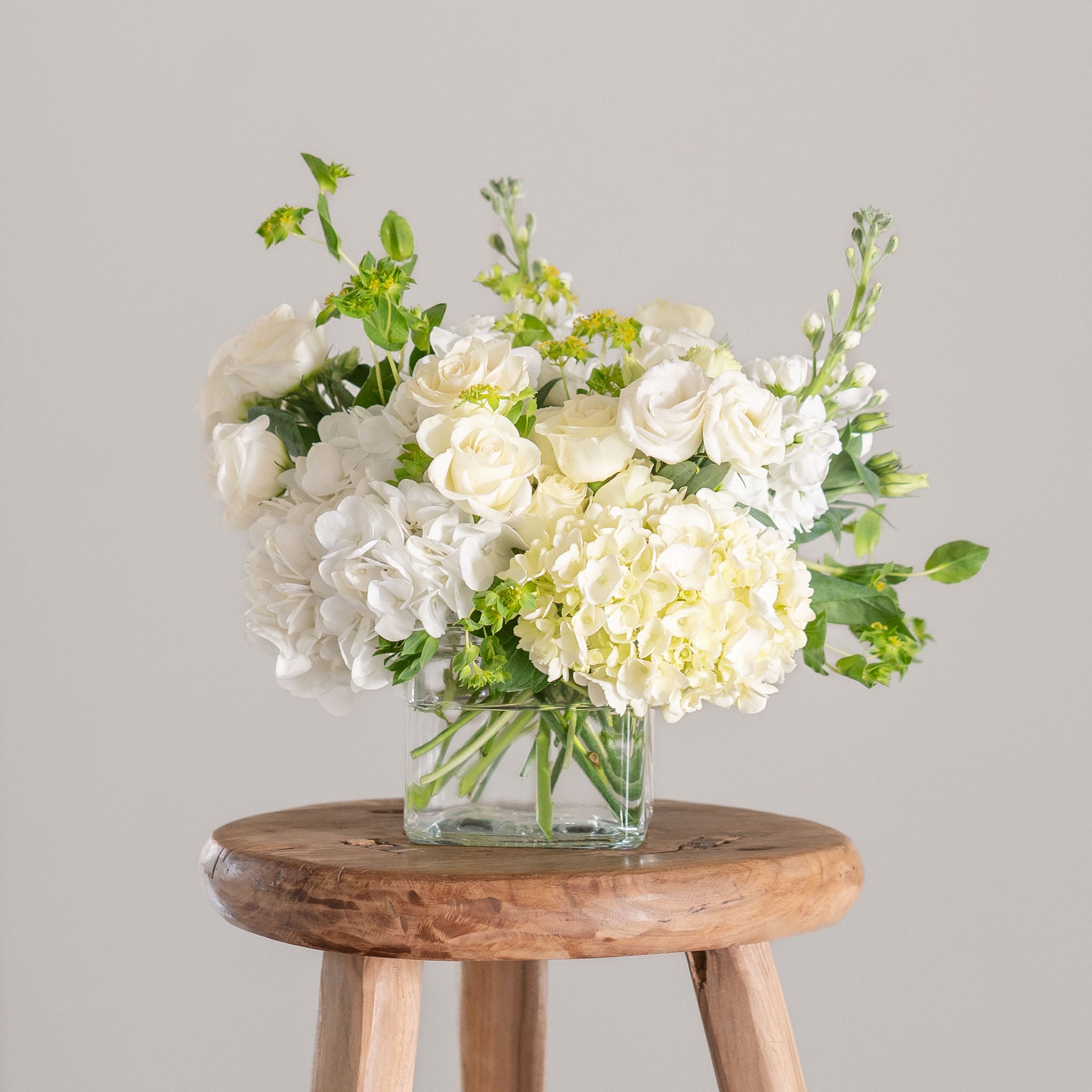 white hydrangea, lisianthus, stock, spray rose and greenery in a clear cube vase on a stool