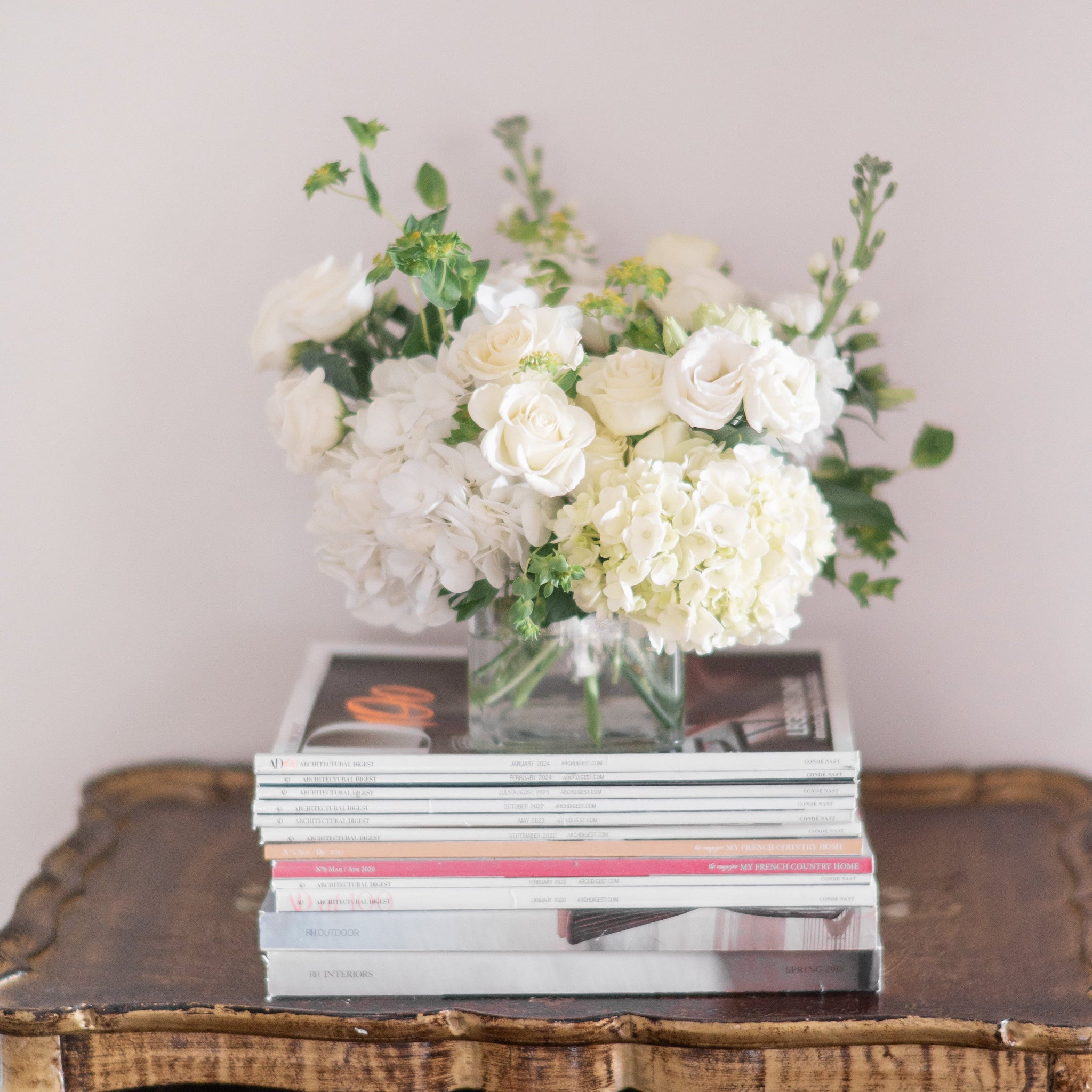 White floral arrangement with white spray roses, white hydrangea, lisianthus and greenery on a stack of books with a wooden table and light background.