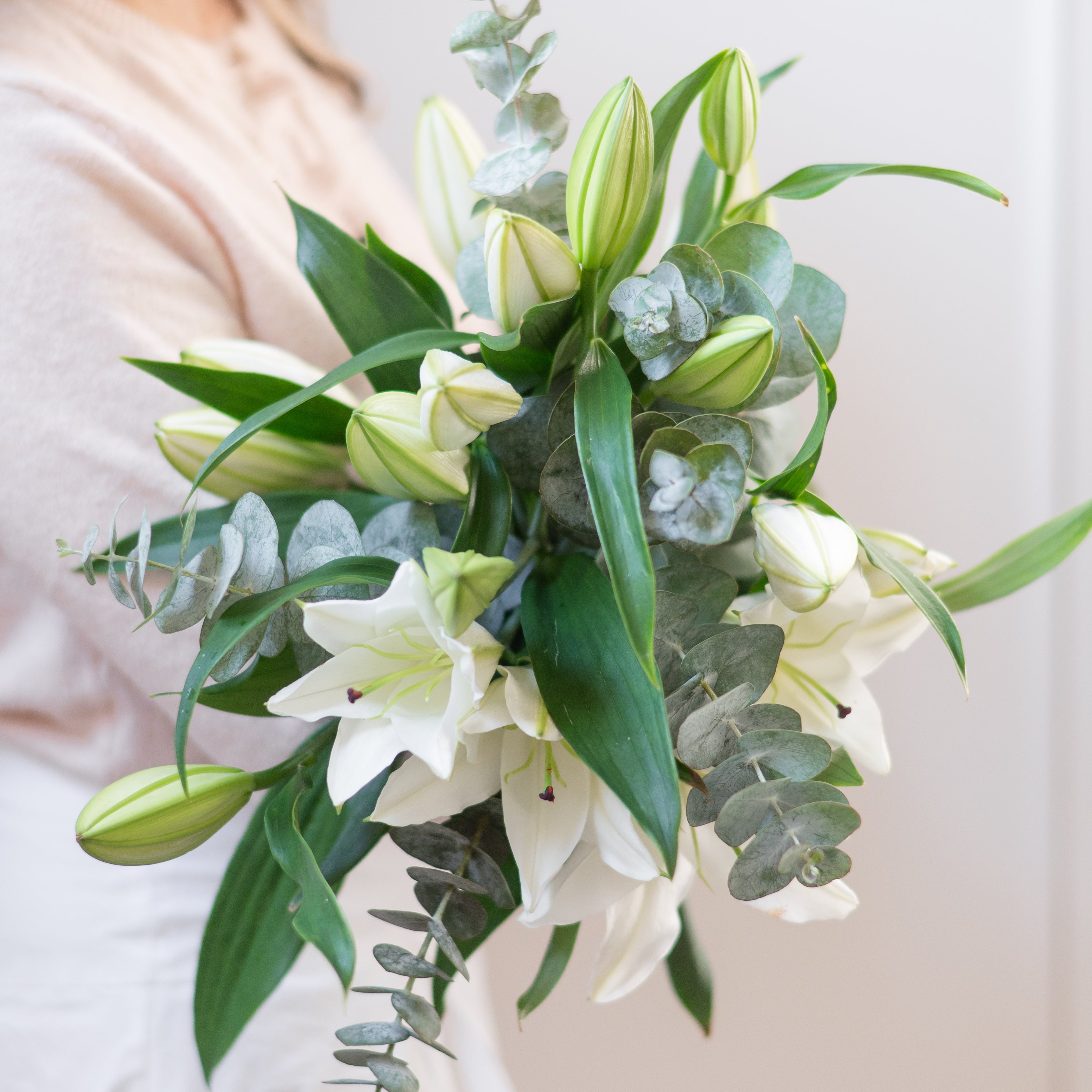 Bouquet of white lilies and greenery held by a person in a light-colored garment.