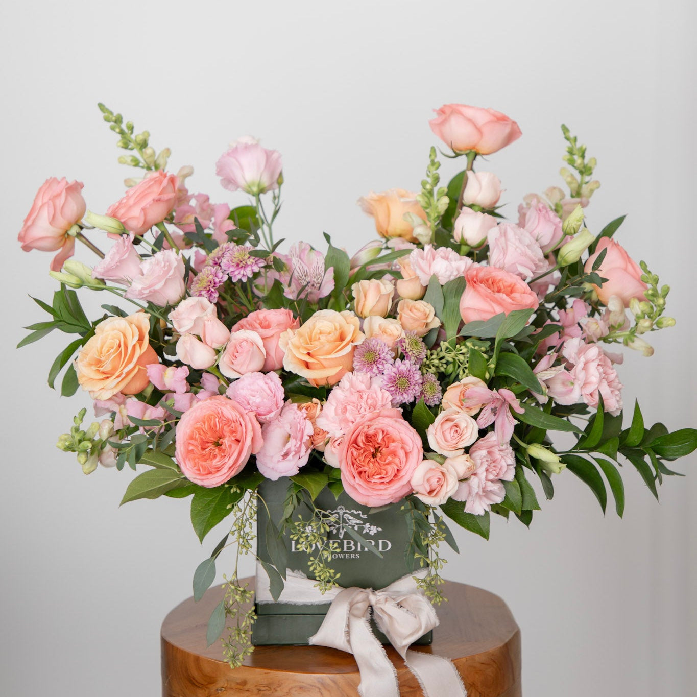 Bouquet of pink and peach flowers in a green lovebird flowers hat box on a  wooden stool with a white background