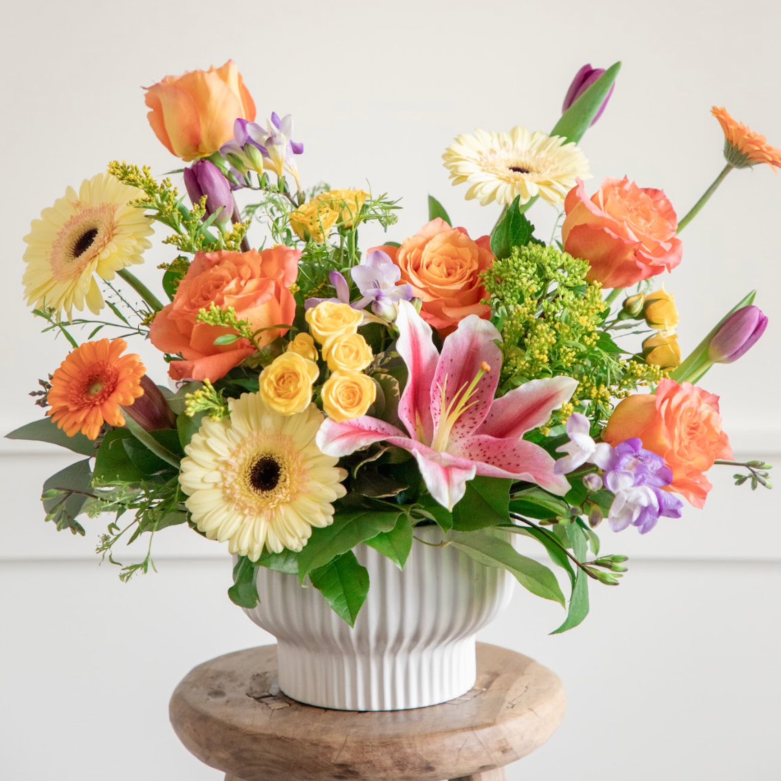 Colorful flower arrangement in a white vase on a wooden stool against a light background