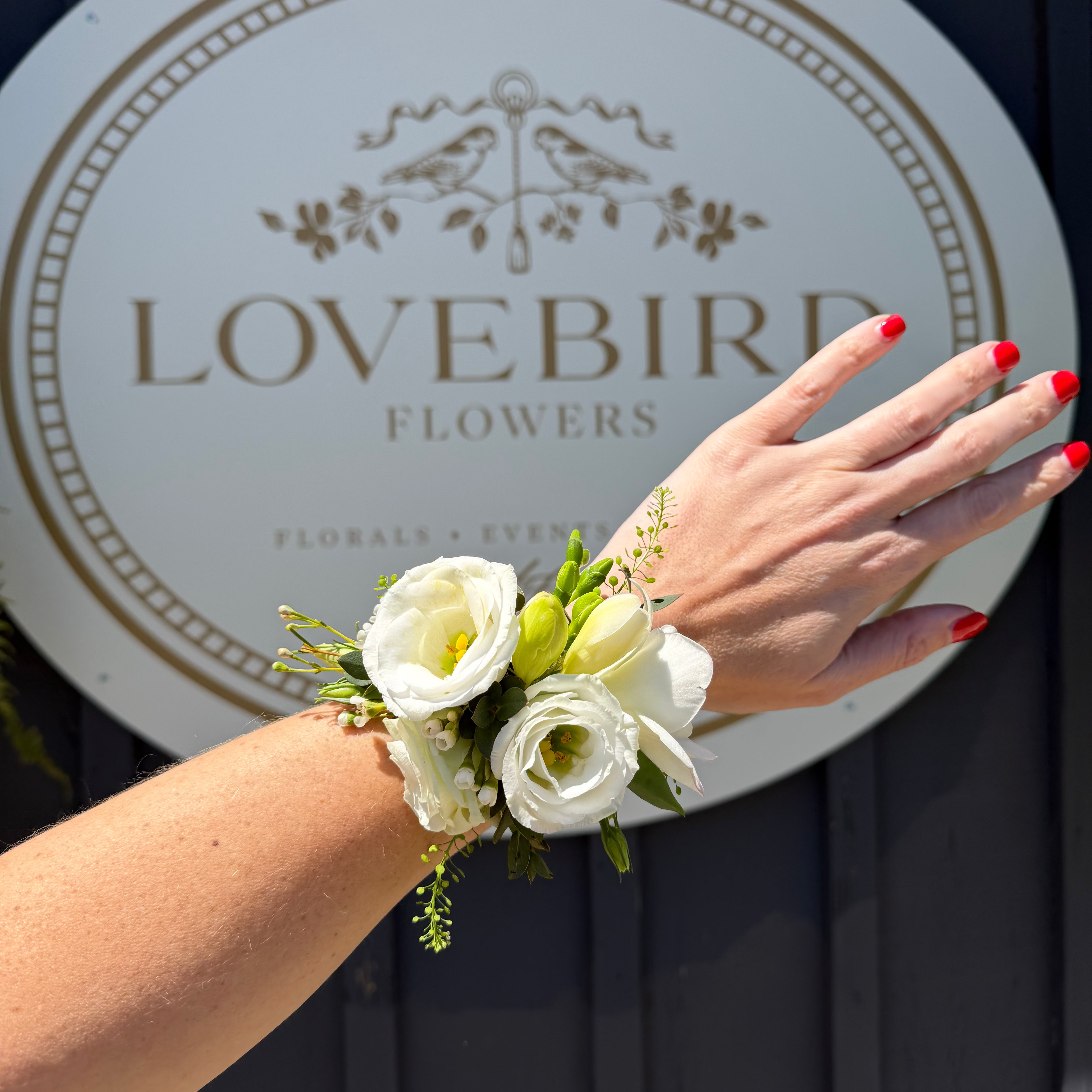 Hand wearing a floral wrist corsage with 'Lovebird Flowers' sign in the background