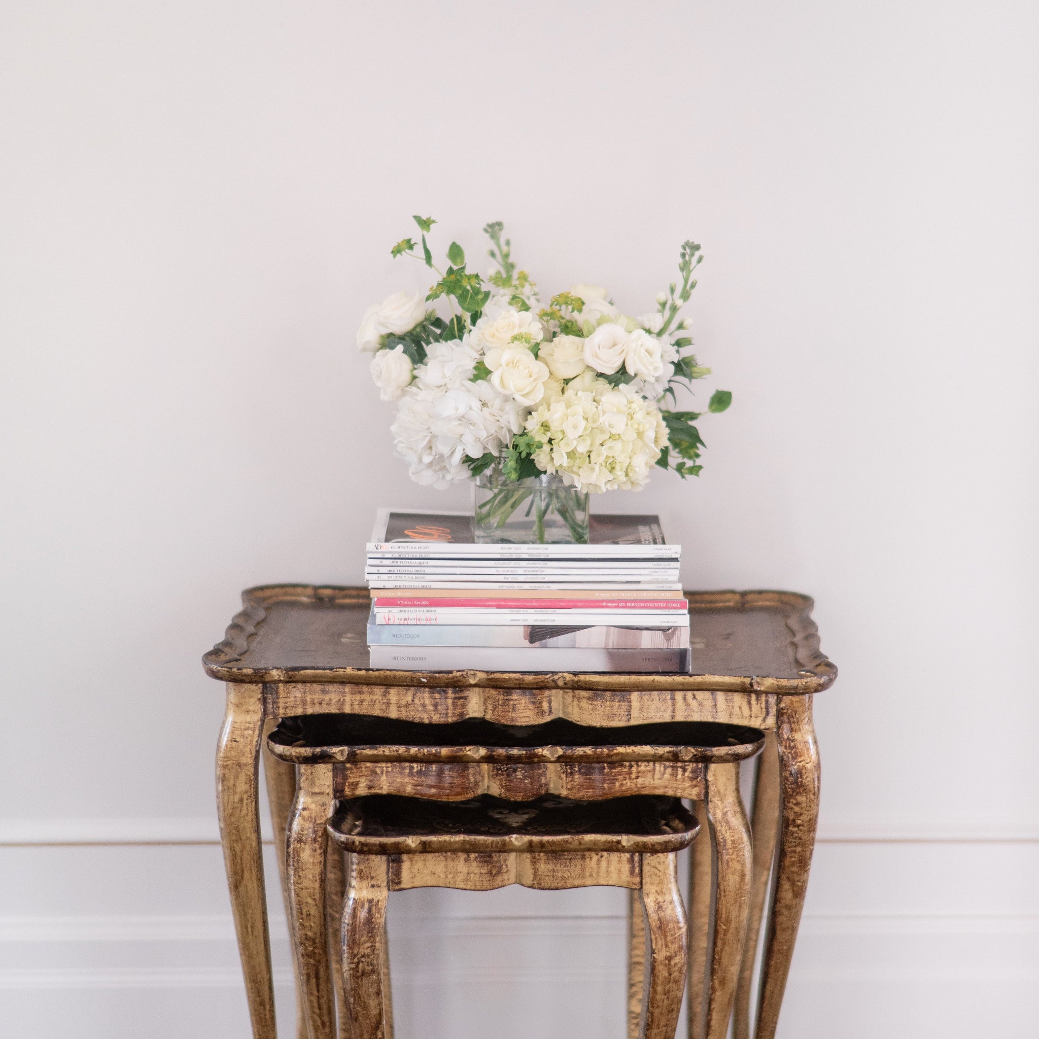 Wooden side table with a vase of white hydrangea, roses, lisianthus, stock and greenery on top of books against a white wall.