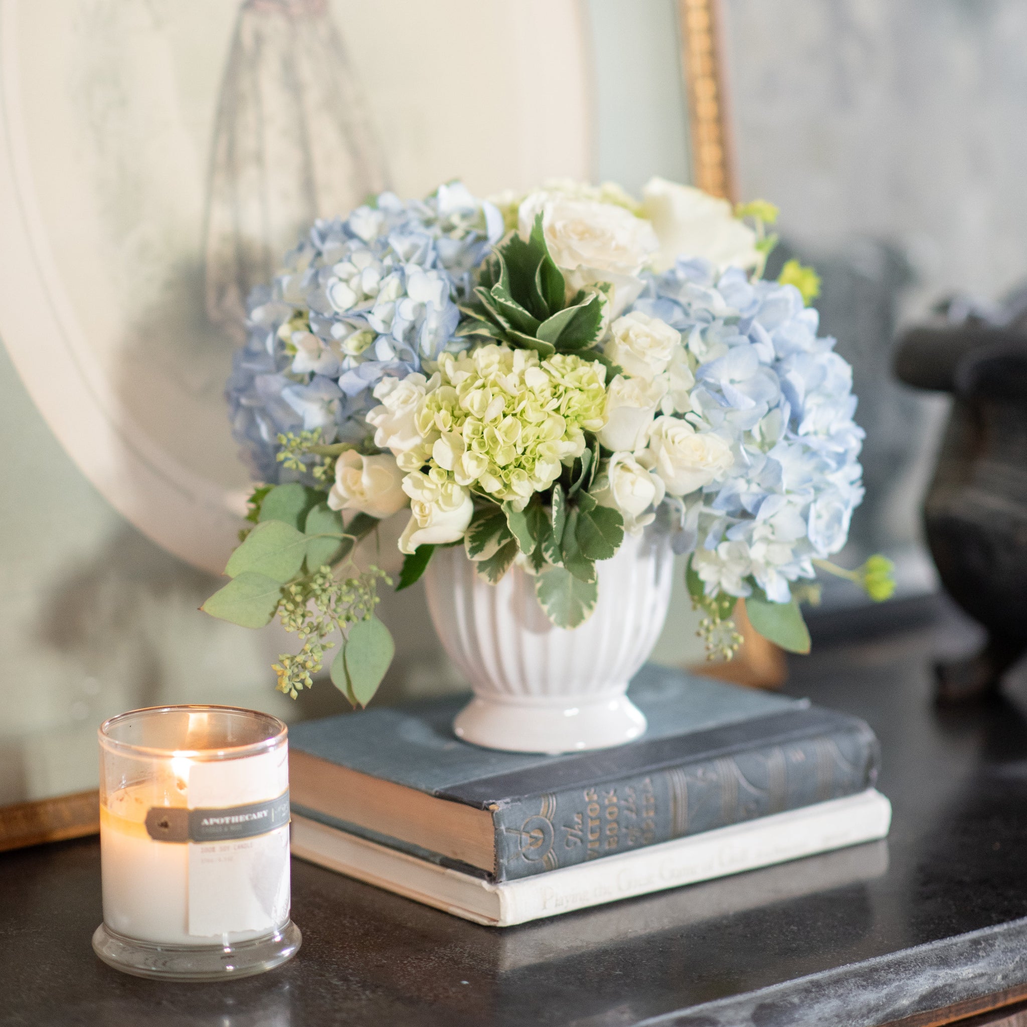 white and blue hydrangeas with white roses in white compote footed vase on old books 