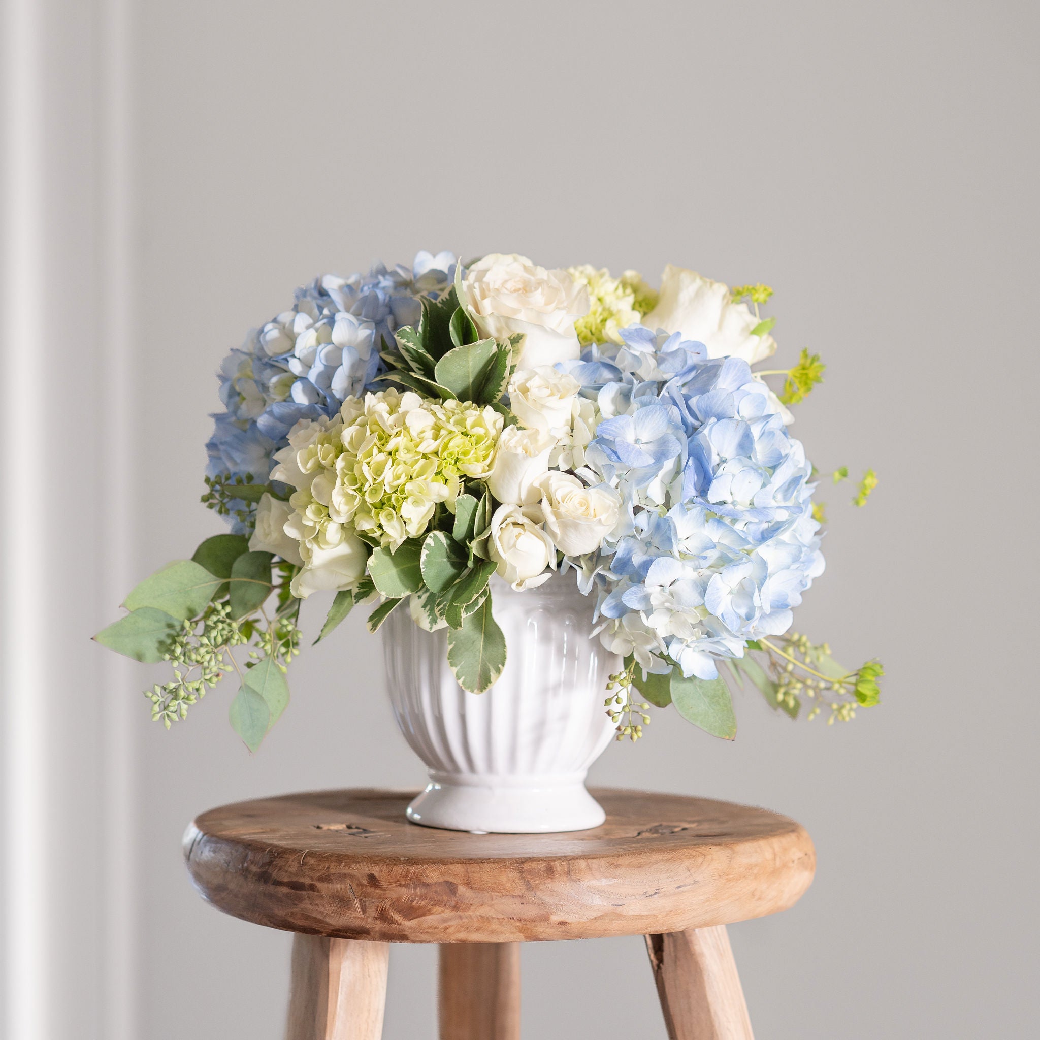 Bouquet of blue, white, and green hydrangeas in a white vase on a wooden stool against a gray background