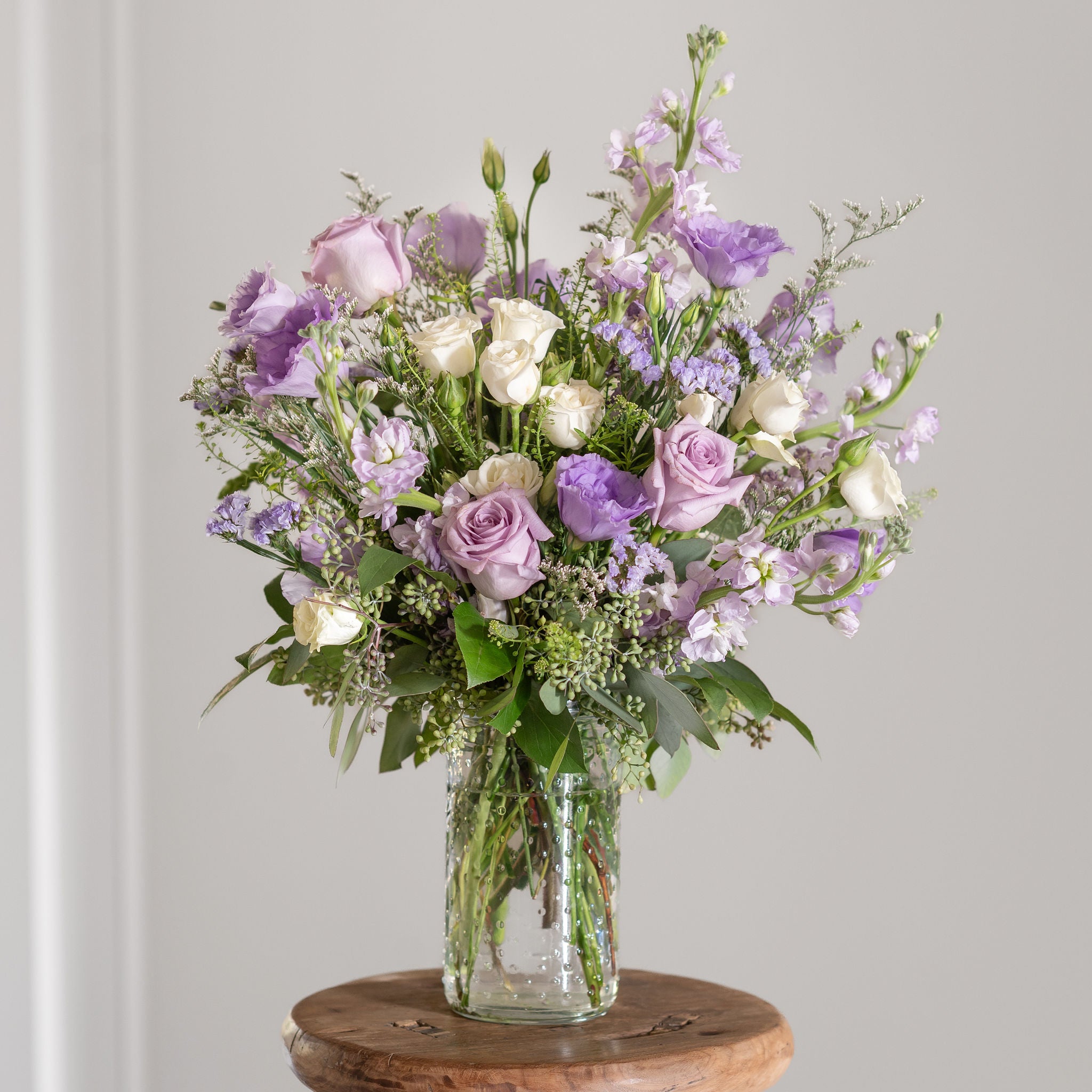Bouquet of purple roses, lavender statice, white spray roses, stock and white flowers in a clear vase on a wooden stand against a gray background