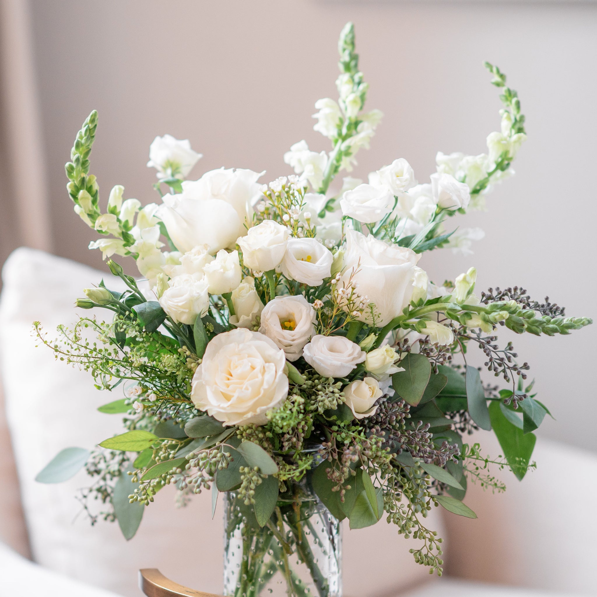 Bouquet of white roses, snap dragons, white lisianthus, seeded eucalyptus and green flowers in a vase on a light-colored surface.