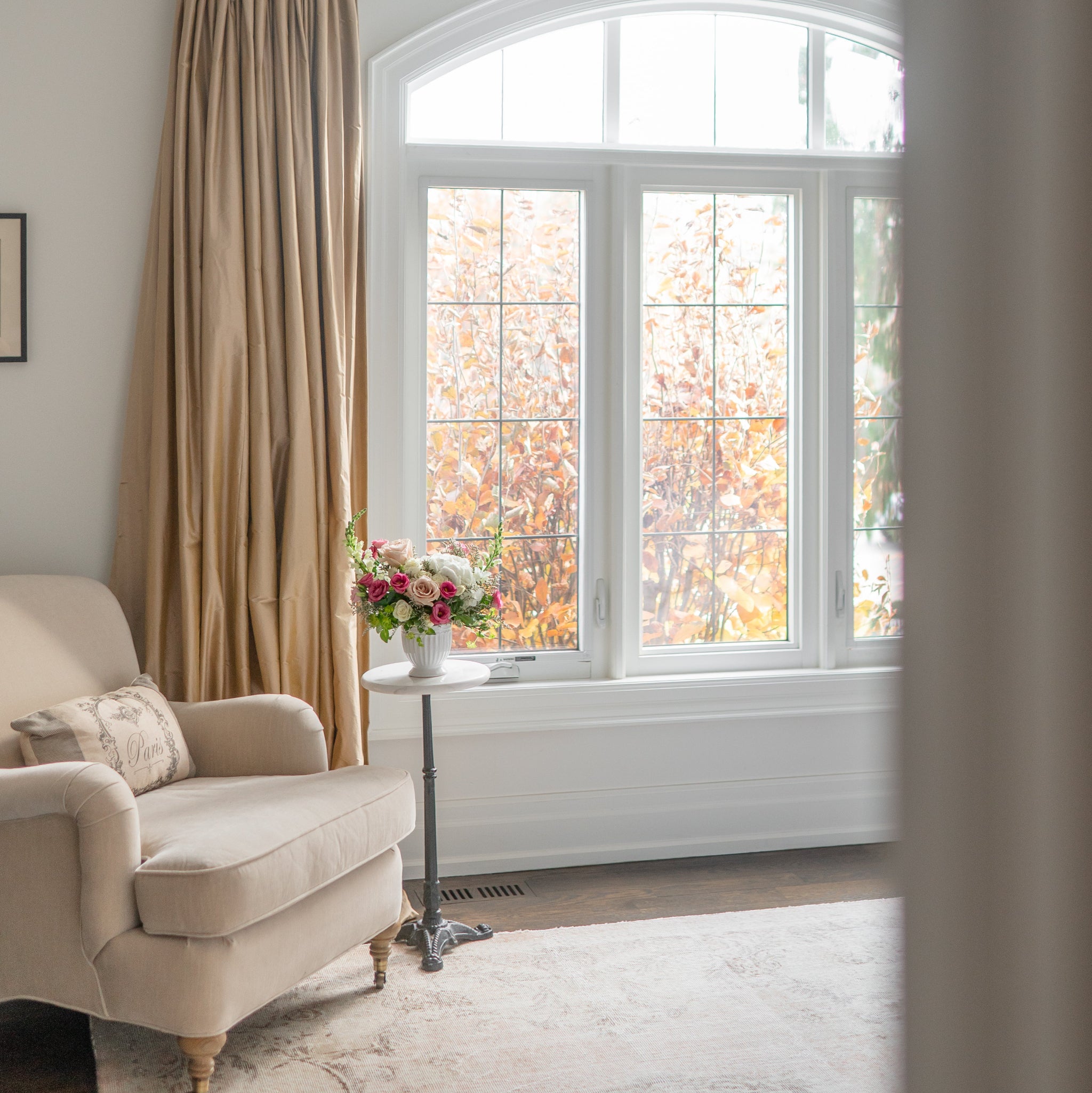 Living room with beige armchair, and white and pink floral arrangement in a white compote vase on a marble side table. 