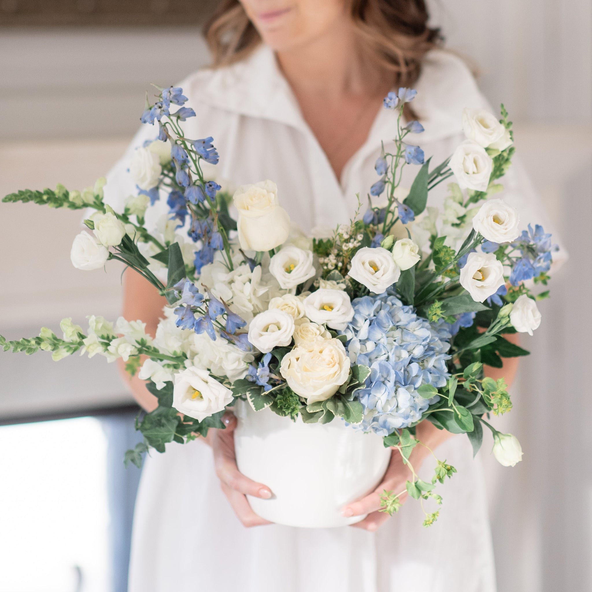 Woman holding a large arrangement of blue delphinium, blue hydrangea, white roses, white lisianthus flowers indoors