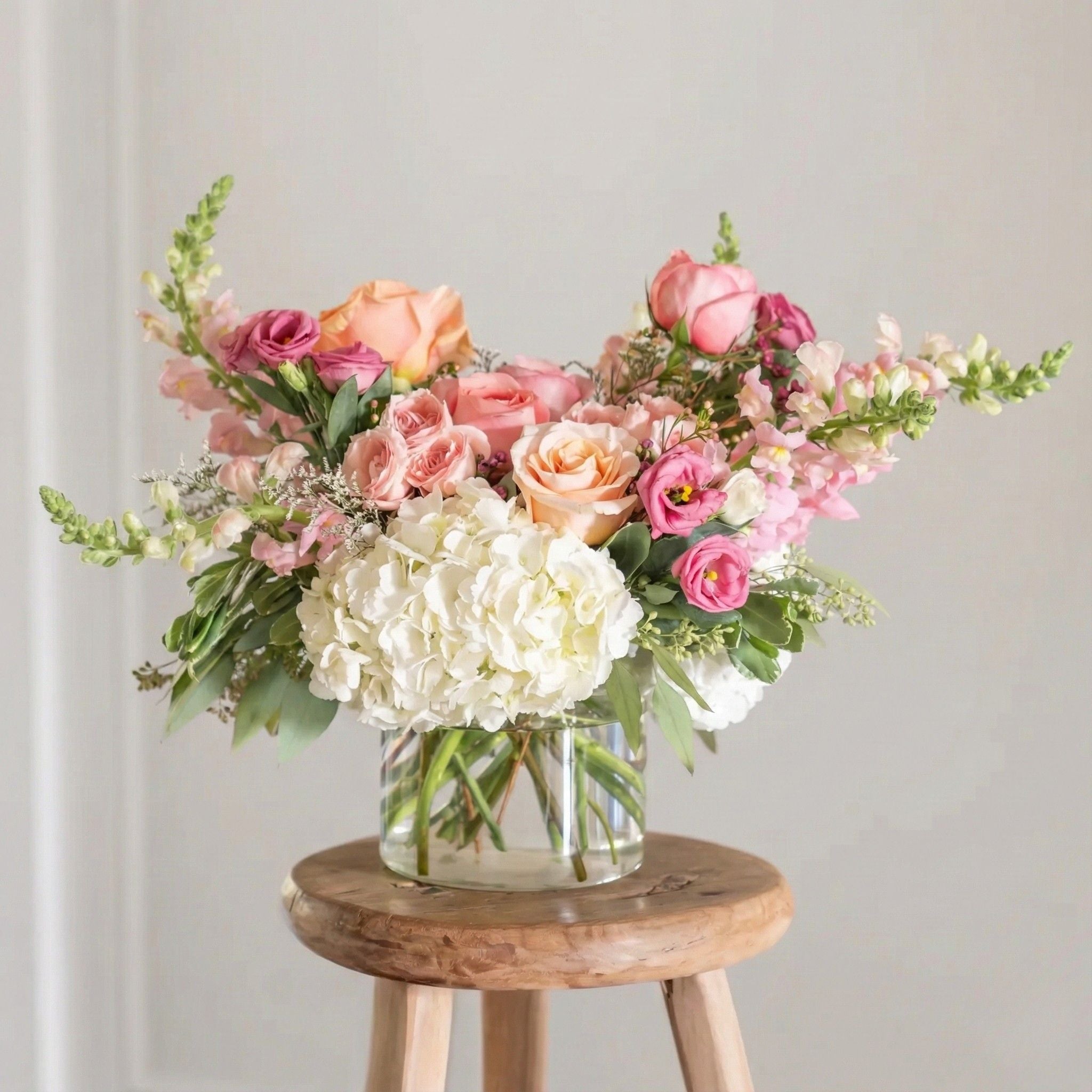 Bouquet of pink and white flowers from Lovebird Flowers in a clear vase on a wooden stool against a gray background