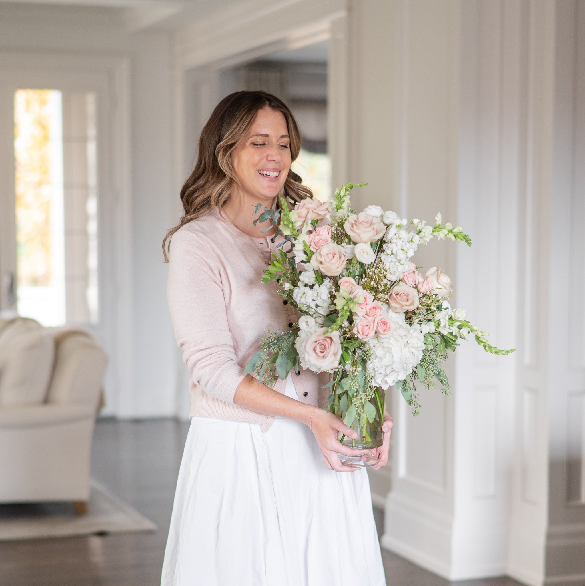 woman walking with a luxurious bouquet of blush and white roses, snapdragons, hydrangea floral arrangement in a tall pedestal glass