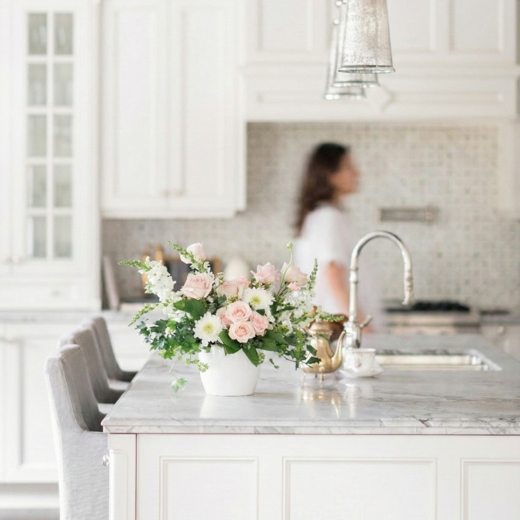 Modern kitchen with marble island, floral arrangement, with blush spray roses, ivey, snap dragons,  and a blurred person in the background.