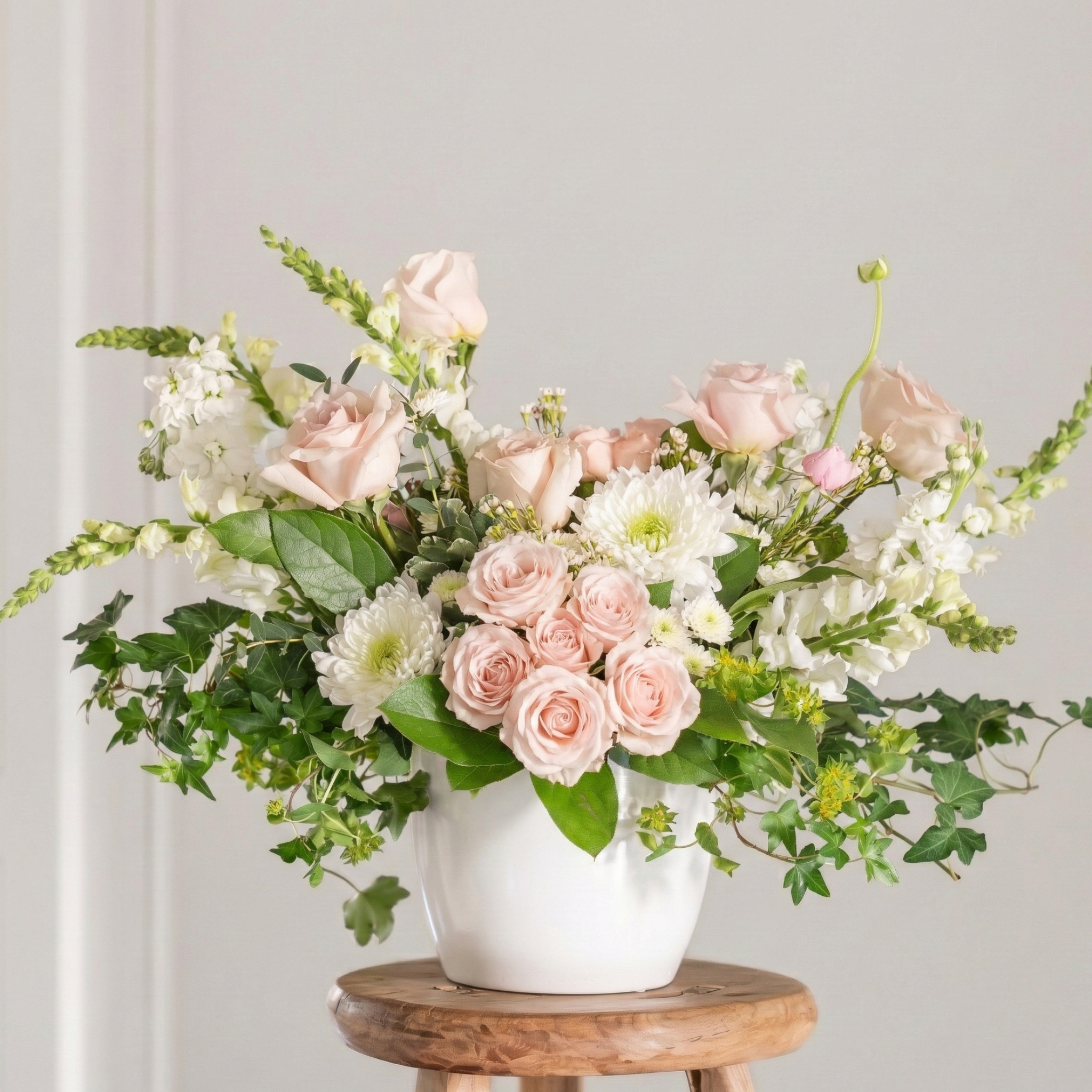 Floral arrangement with pink and white flowers in a white vase on a wooden stool against a light gray background